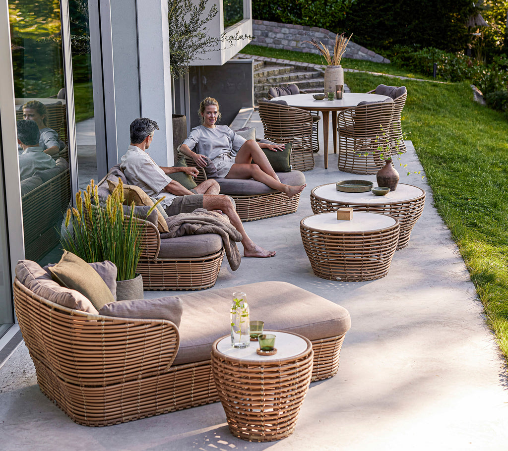 Dos personas se relajan en muebles trenzados rodeados de mesas y vegetación, disfrutando de un ambiente relajado al aire libre.