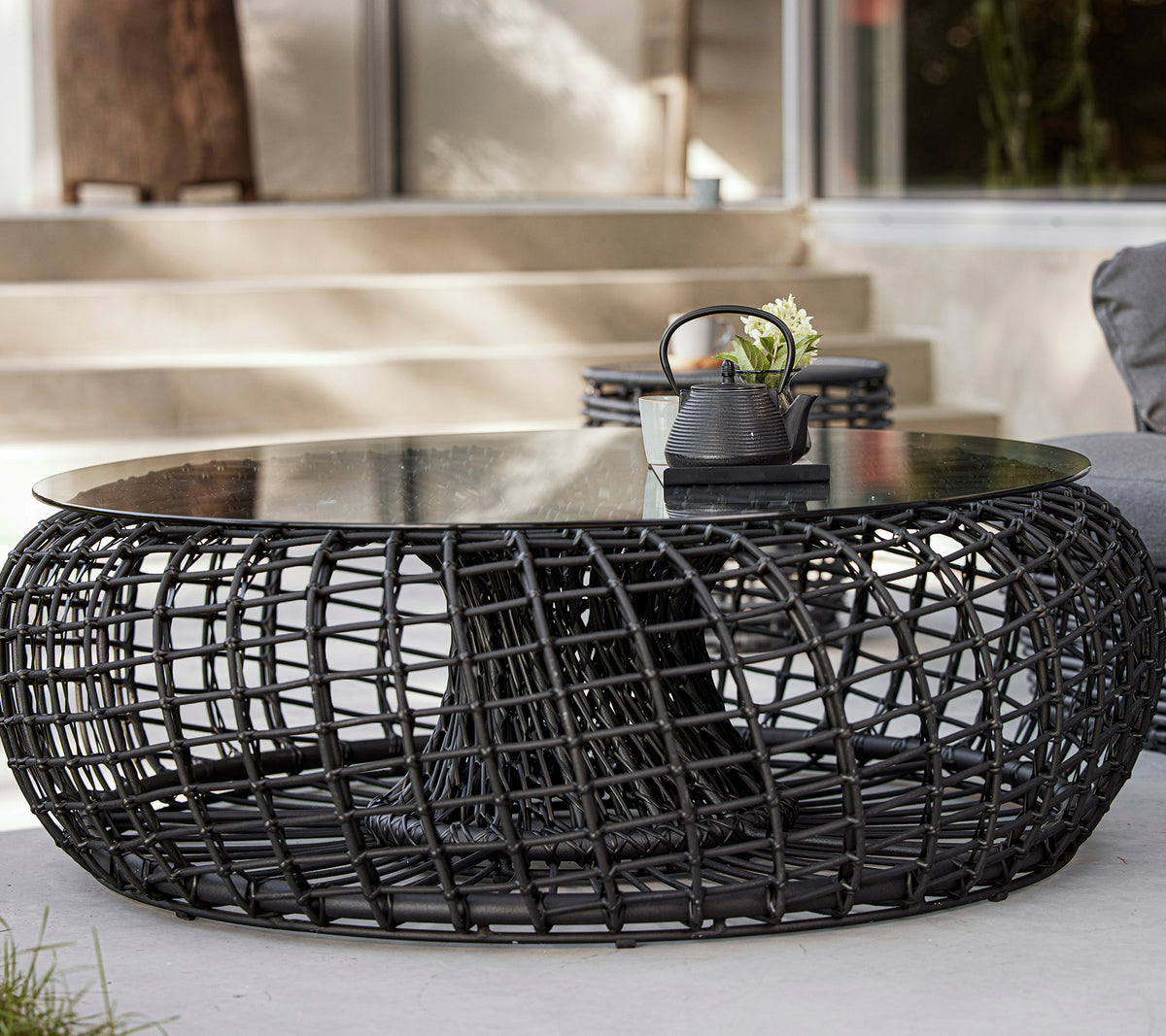Una mesa de café moderna y redonda con una base negra trenzada y una tapa de vidrio, que presenta elementos decorativos como una planta y un pequeño tazón.
