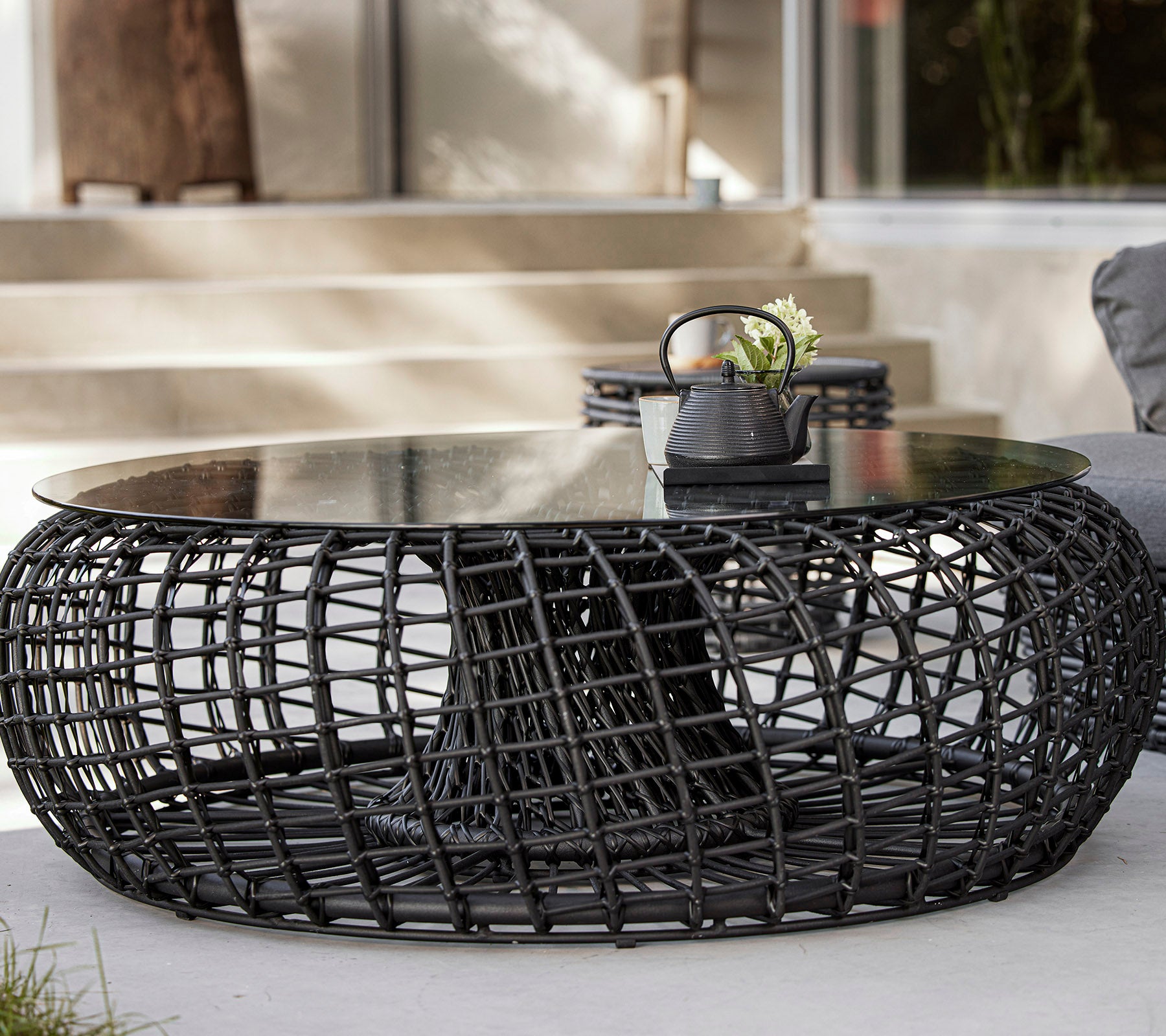 Una mesa de café moderna y redonda con una base negra trenzada y una tapa de vidrio, que presenta elementos decorativos como una planta y un pequeño tazón.