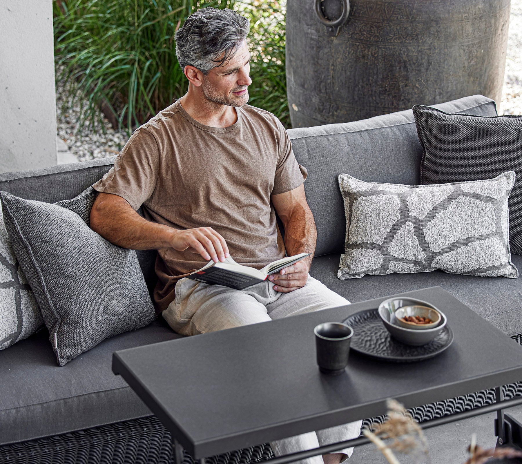 Un hombre está sentado cómodamente en un sofá, leyendo un libro con una taza y un tazón en una mesa de café a su lado, rodeado de cojines decorativos.
