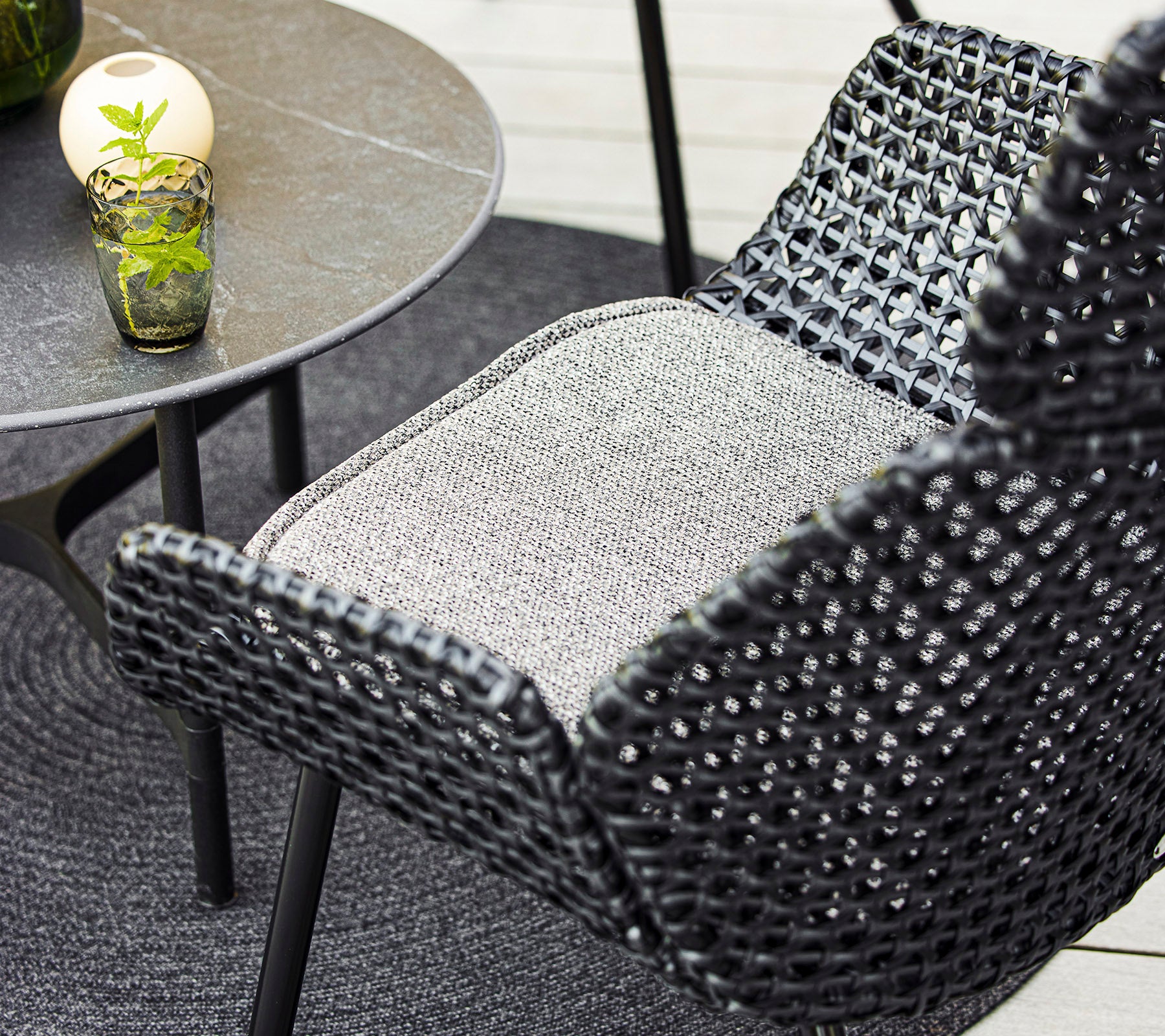 Una vista en primer plano de una elegante silla tejida y una mesa pequeña con un vaso decorativo y una planta, acompañada de una alfombra circular acogedora.