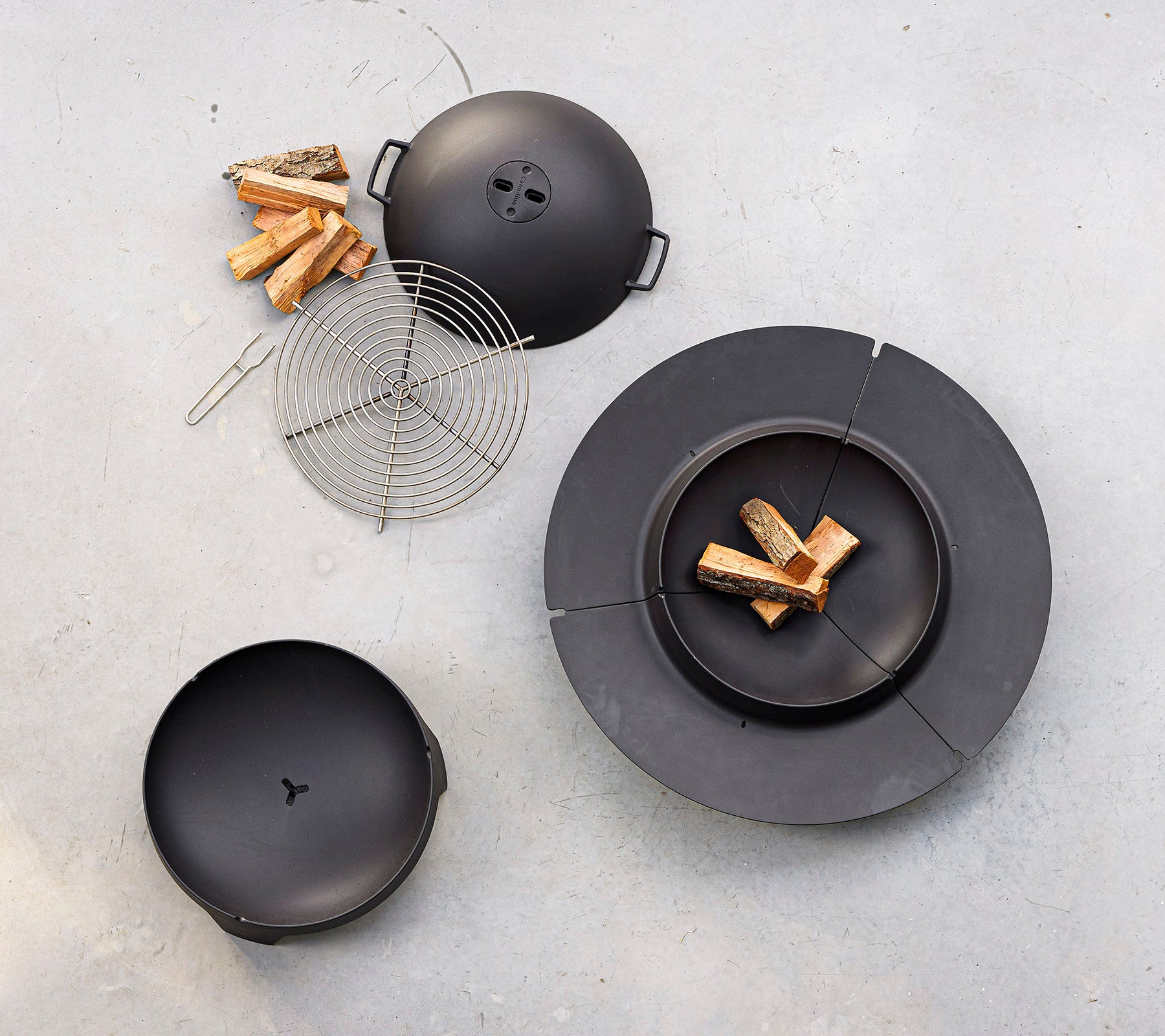 Un juego de cocina negro que incluye una parrilla circular con tapa, una rejilla para asar y piezas de leña dispuestas cerca.