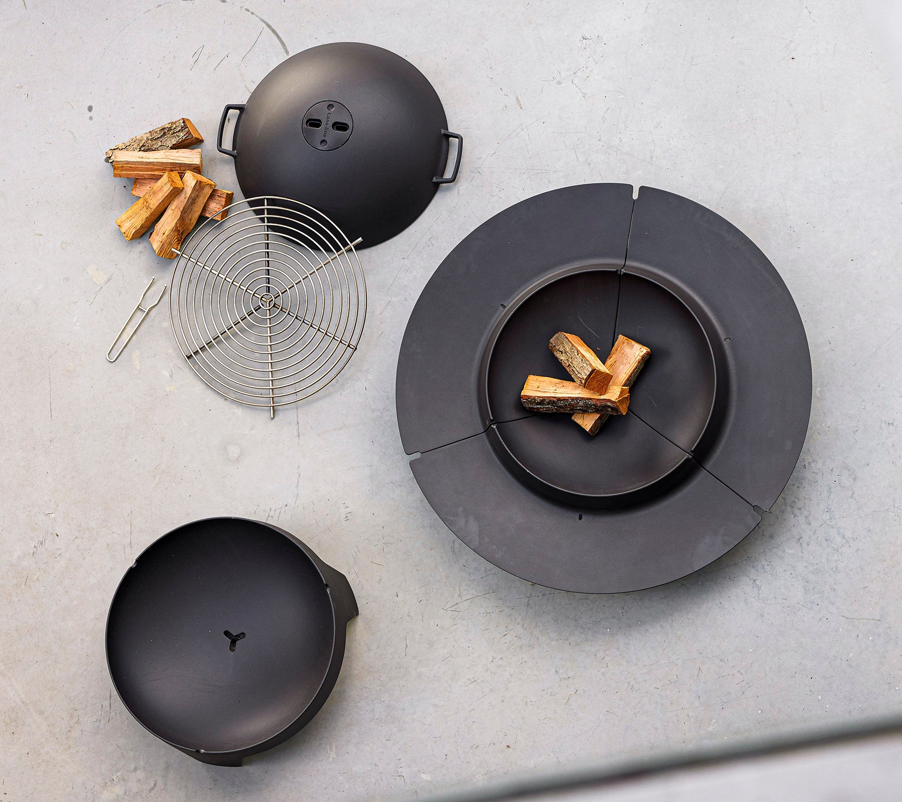 Equipo de cocina negro dispuesto con leña y una rejilla de alambre, diseñado para asar al aire libre y cocinar con fuego.