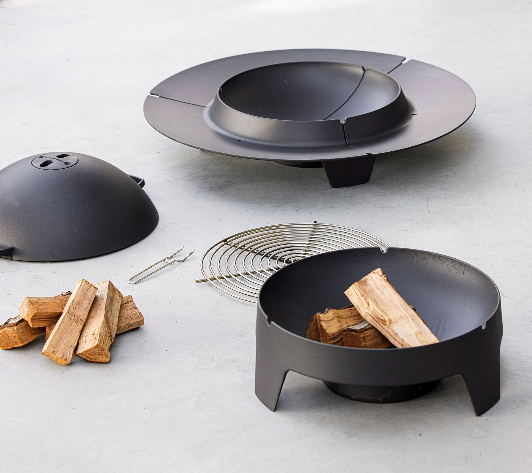 Un conjunto de fogón negro que incluye un cuenco, una tapa, una rejilla y madera apilada, diseñado para reuniones al aire libre.