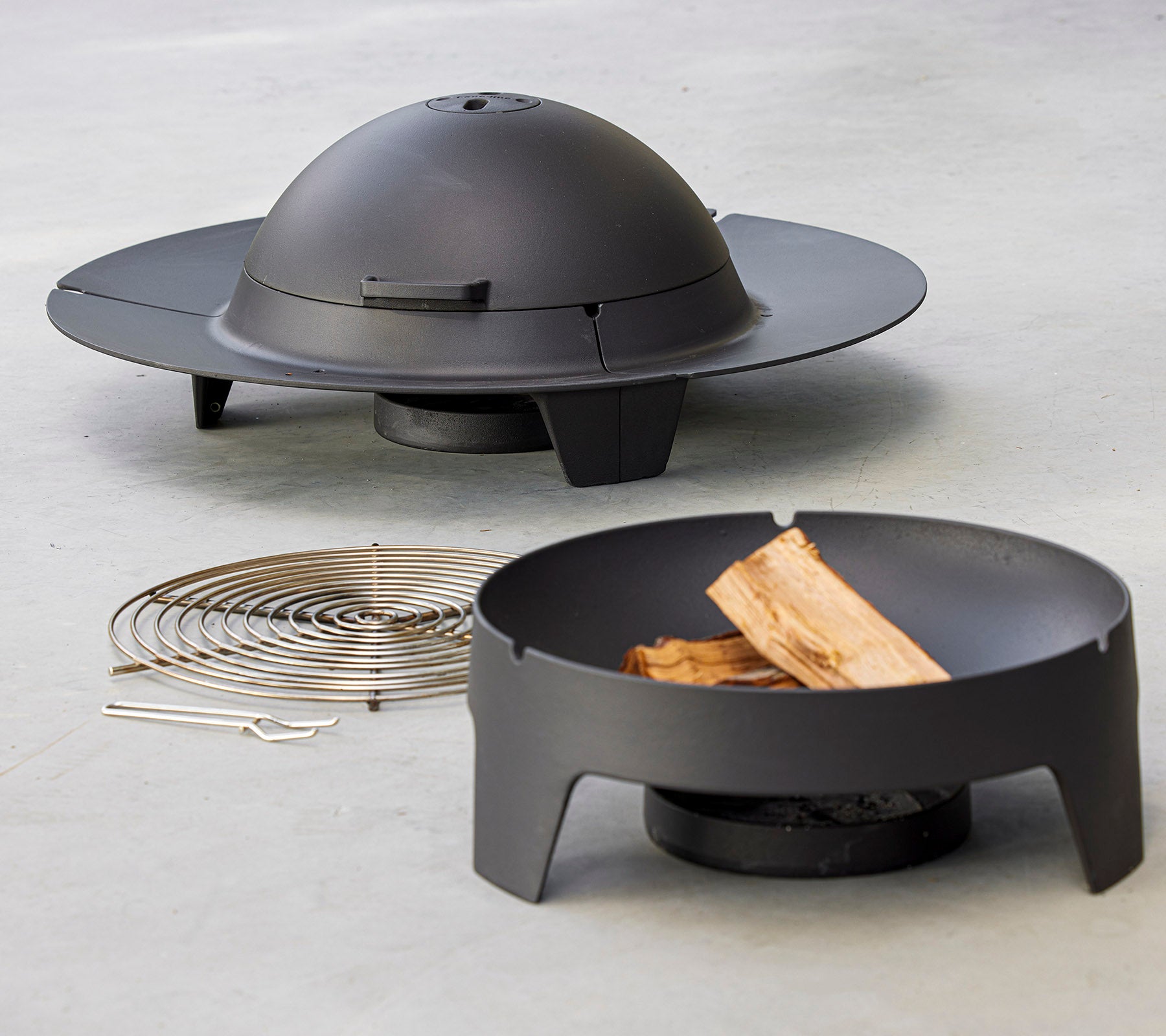 Un juego de fuego moderno con un diseño circular elegante y una tapa de cúpula, acompañado de una rejilla de parrilla y trozos de madera.