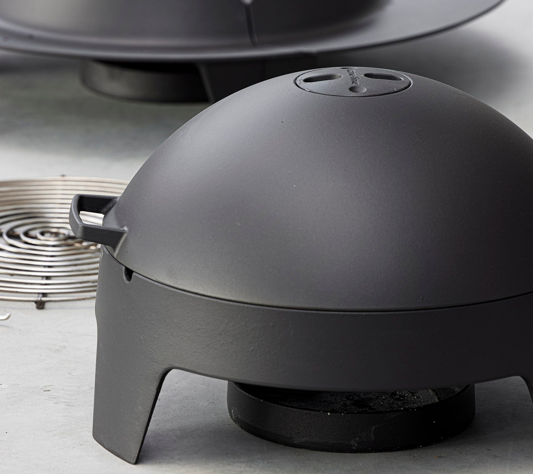 Aparato de cocina negro mate con una tapa en forma de cúpula y una fuente de calor extraíble, diseñado para asar a la parrilla y hacer barbacoas al aire libre.