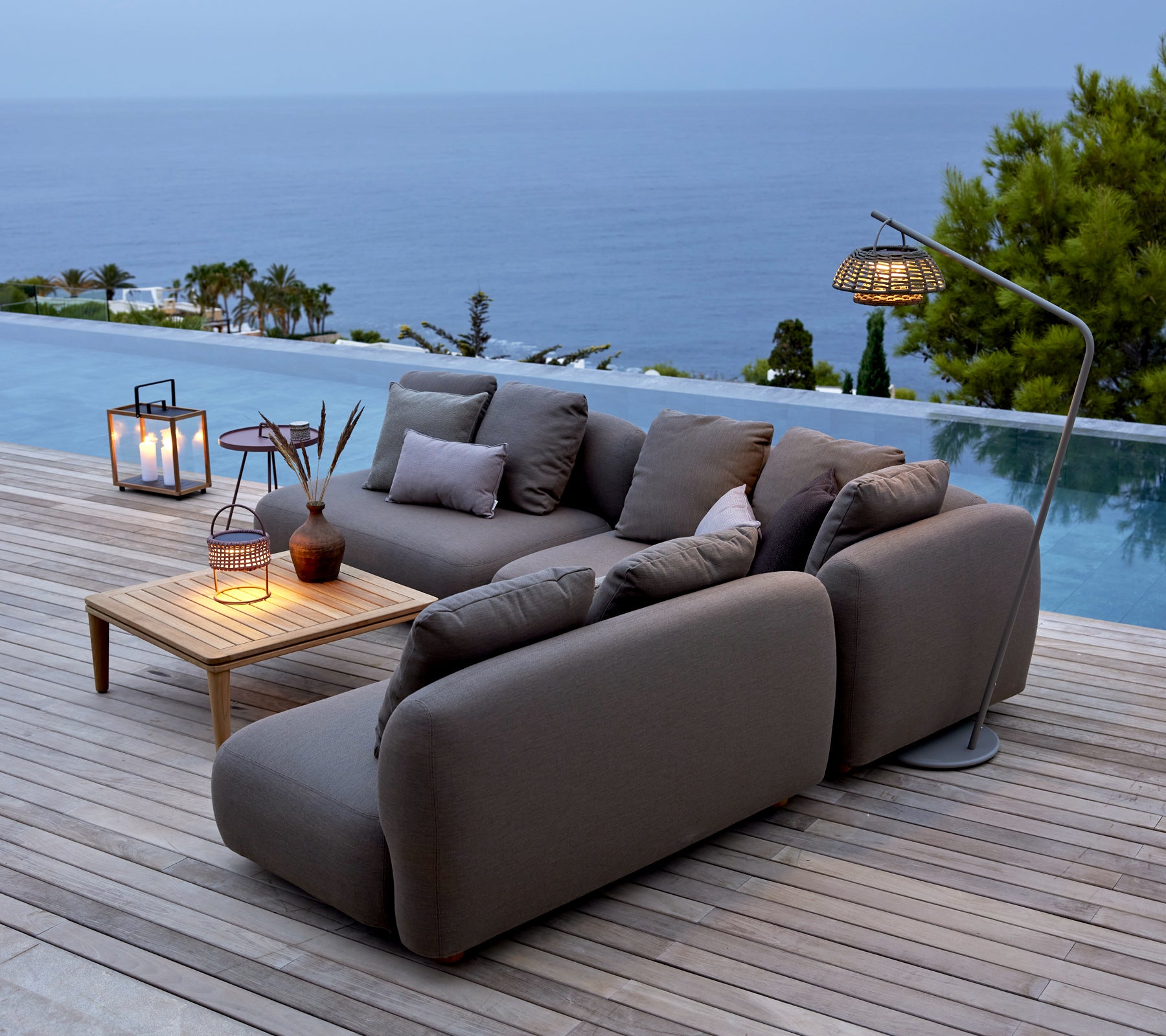 Sofá gris con mesa de madera y faroles junto a una piscina con vista al océano.