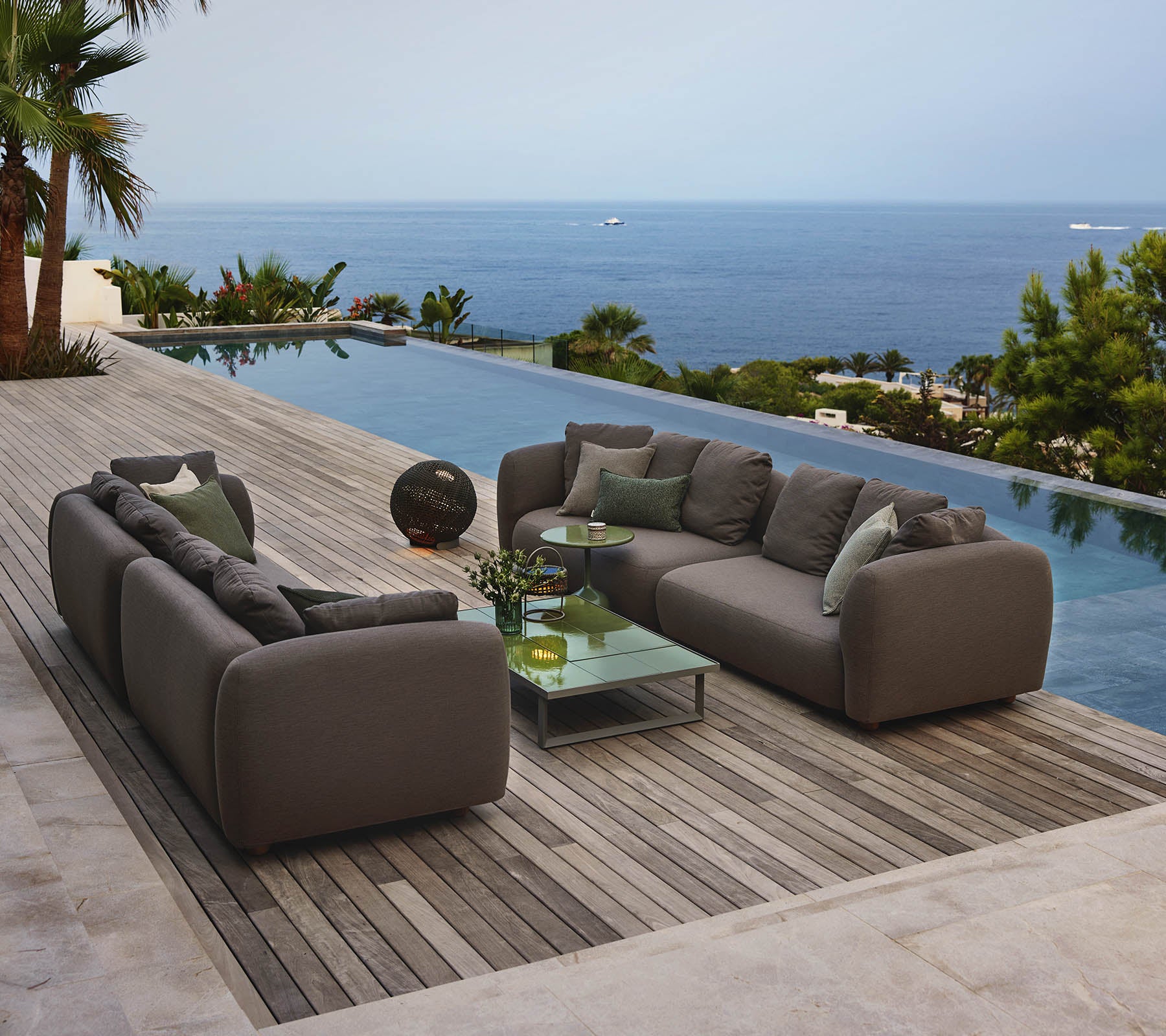 Asientos de lounge modernos dispuestos alrededor de una mesa de café de vidrio, con vista a una serena piscina y una amplia vista al océano, rodeados de vegetación.
