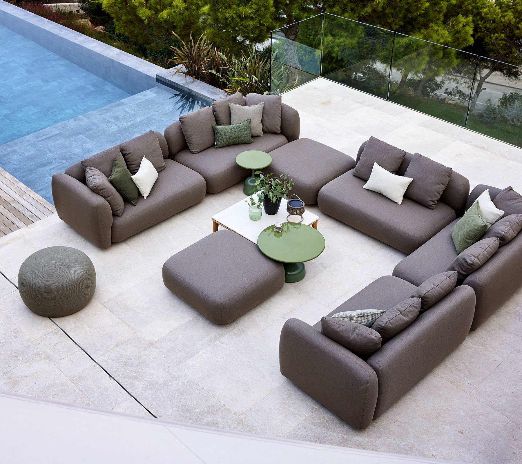 Un diseño moderno de salón con un sofá seccional, un otomán redondo y mesas auxiliares verdes, rodeado de vegetación y una piscina tranquila.