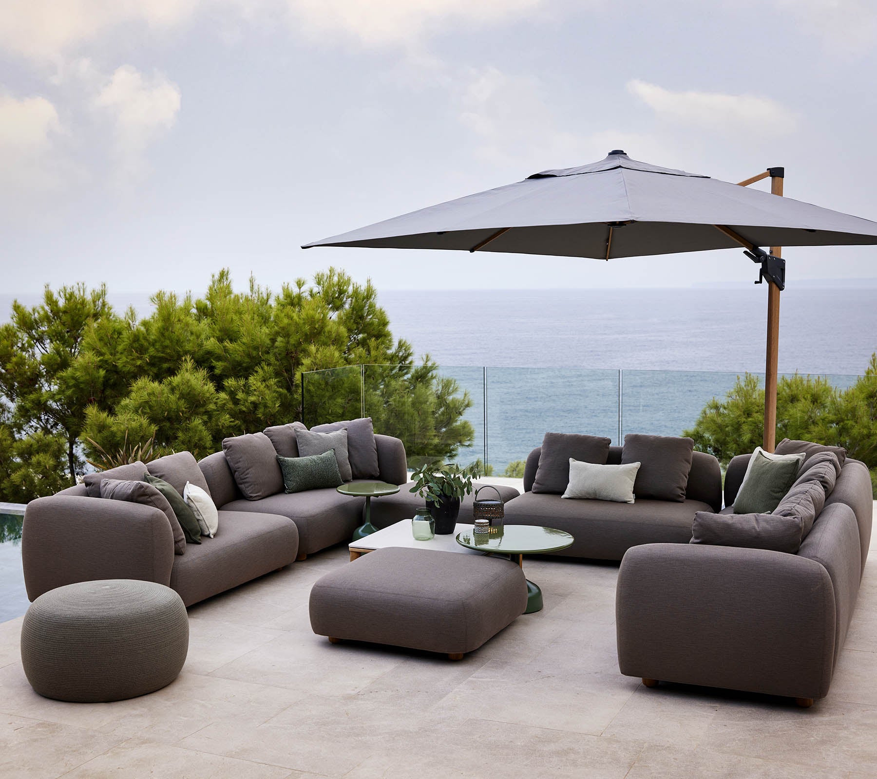 Conjunto de asientos moderno con cojines suaves, acompañado de una mesa redonda y un gran parasol, rodeado de vegetación y una vista escénica.