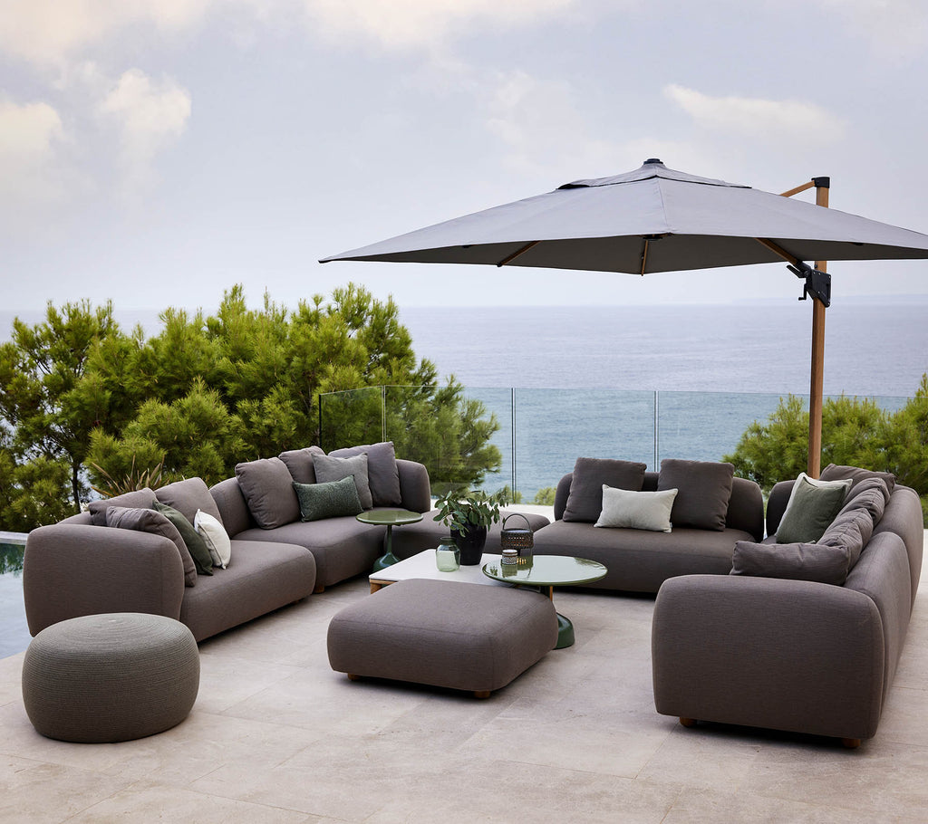 Conjunto de asientos moderno con cojines suaves, acompañado de una mesa redonda y un gran parasol, rodeado de vegetación y una vista escénica.