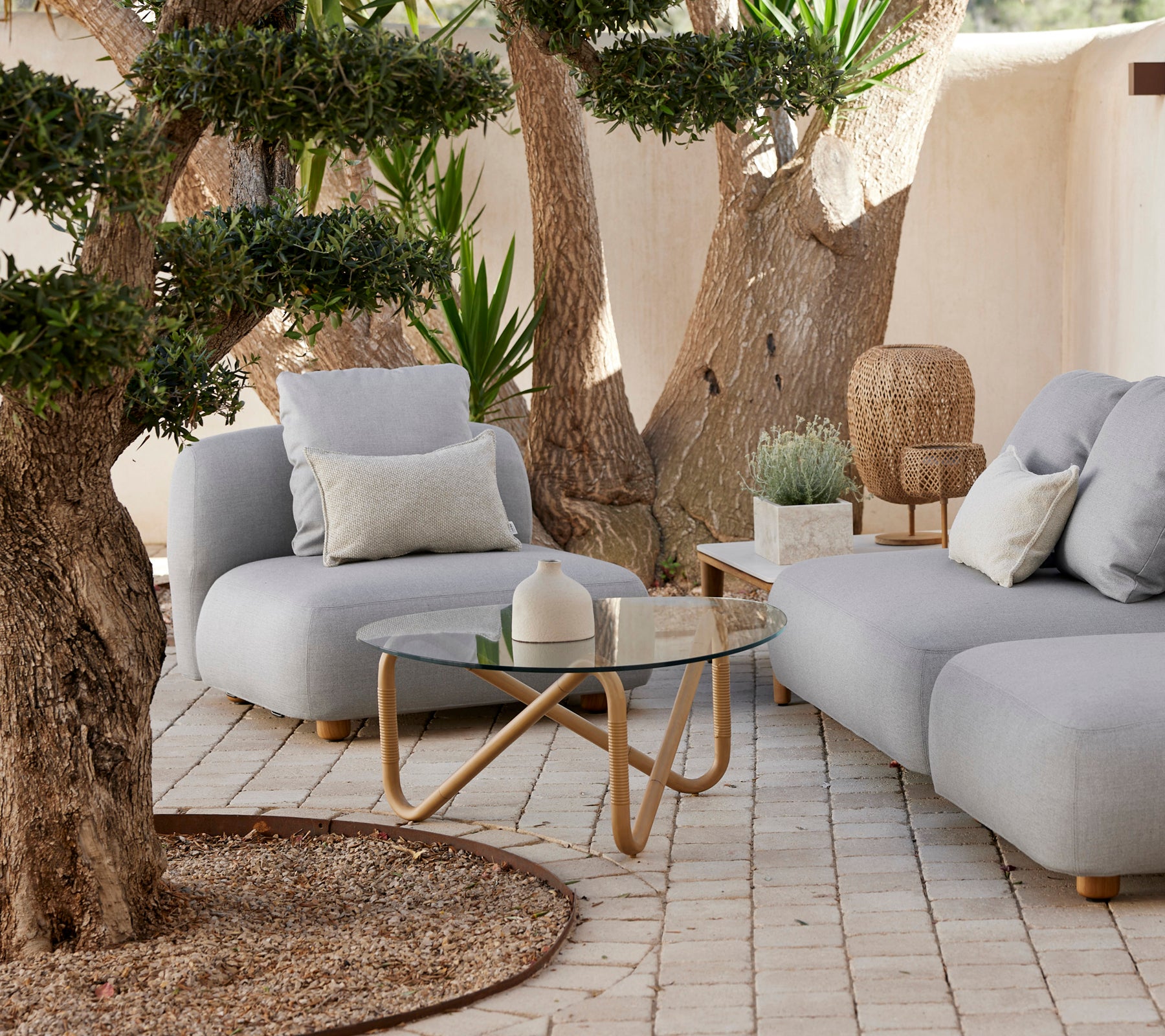 Sofá gris moderno con cojines y una mesa de café de vidrio en un ambiente exterior relajado.