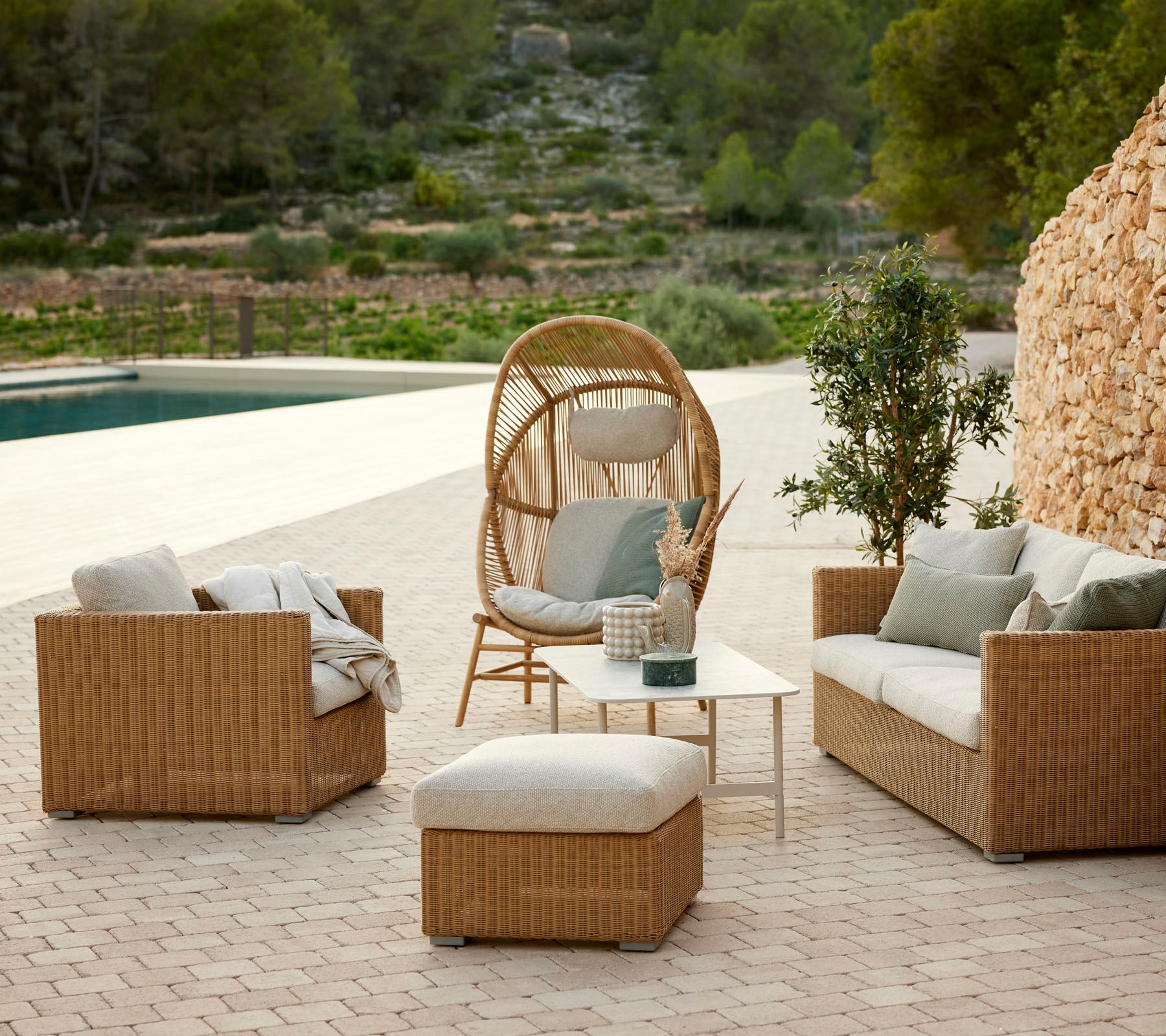 Agradable disposición de asientos al aire libre con sillones de mimbre, una silla colgante circular, un ottomán y una mesa pequeña entre la vegetación.