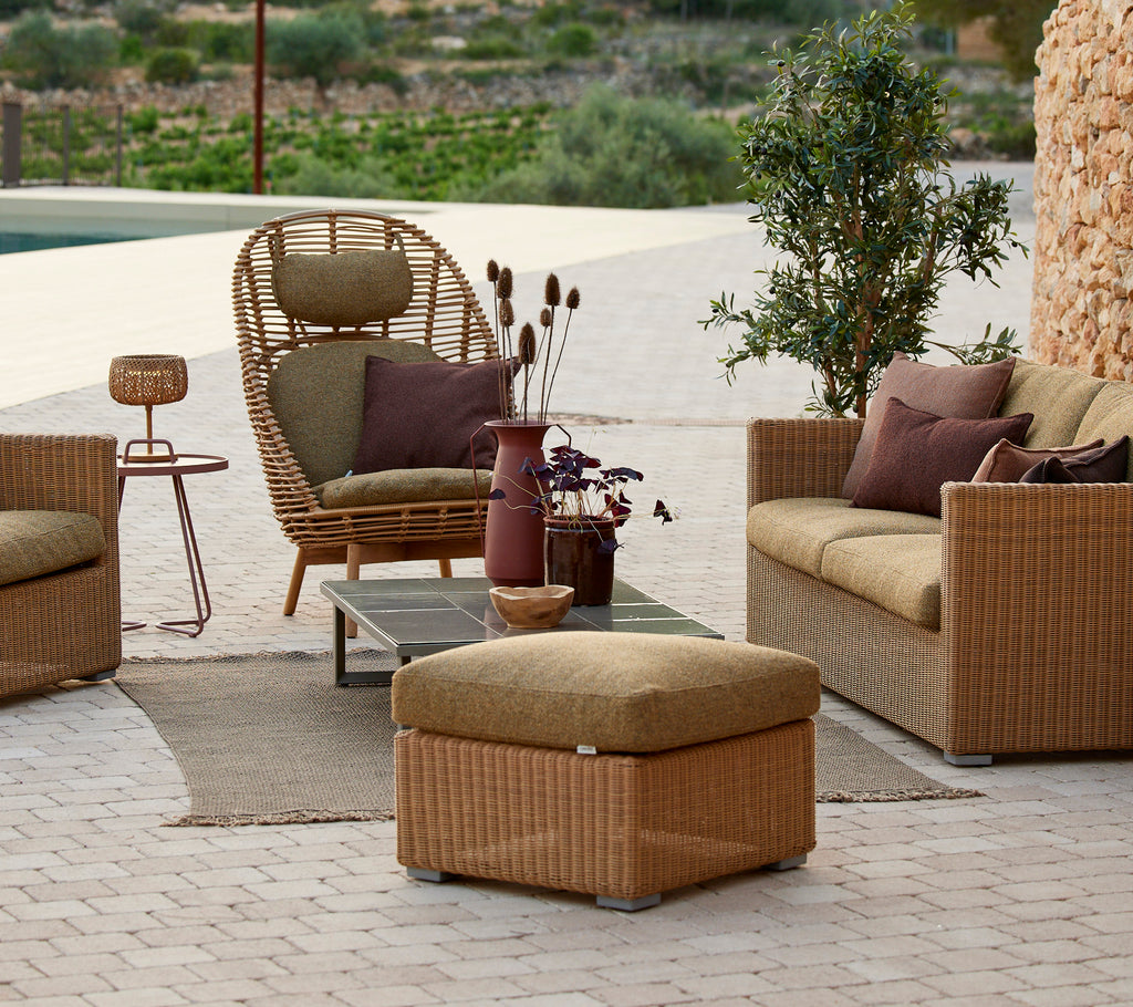Área de lounge con silla de ratán, sofá beige y mesa auxiliar con un jarrón morado.