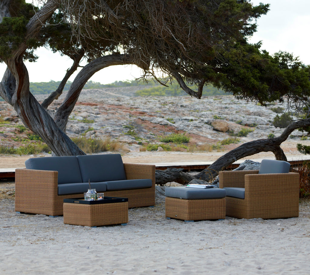 Muebles de salón modernos en marrón con cojines grises situados en un entorno natural al aire libre.