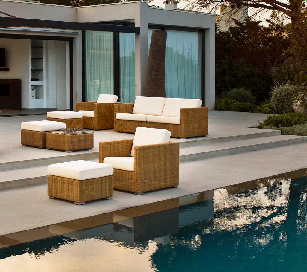 Área moderna de salón al aire libre con sofá marrón, sillas y cojines junto a una piscina.