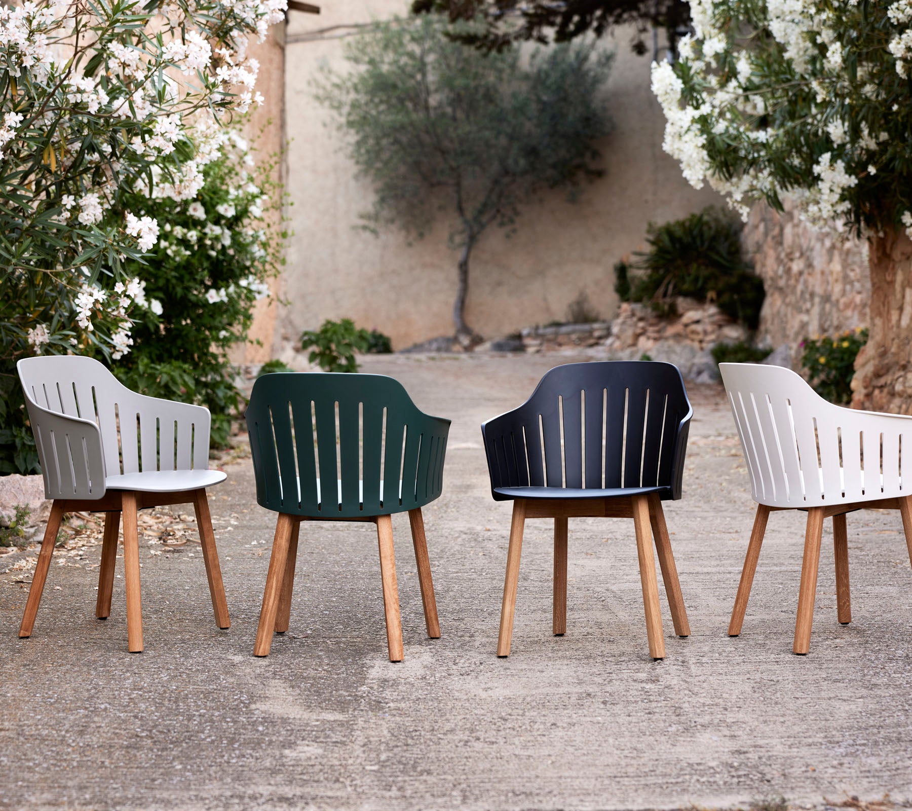 Cuatro sillas elegantes en diferentes colores con patas de madera, mostrando un diseño moderno y respaldos de lamas entre la vegetación.