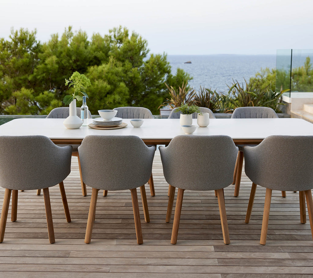 Una mesa moderna con sillas tapizadas en gris, rodeada de vegetación y con una vista serena del océano, perfecta para cenar y reuniones.