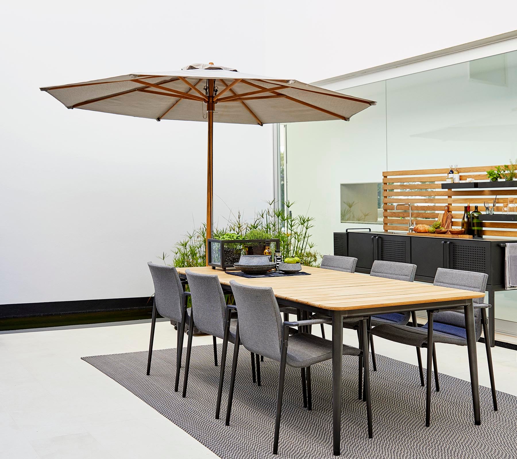 Un conjunto de comedor clásico con una mesa de madera con una sombrilla y sillas grises, rodeado de plantas decorativas sobre una alfombra elegante.