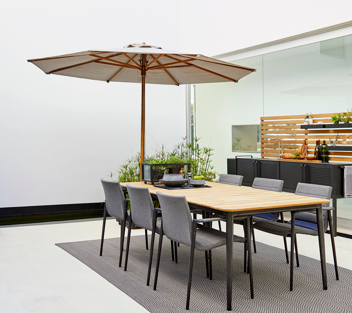 Un conjunto de comedor clásico con una mesa de madera con una sombrilla y sillas grises, rodeado de plantas decorativas sobre una alfombra elegante.