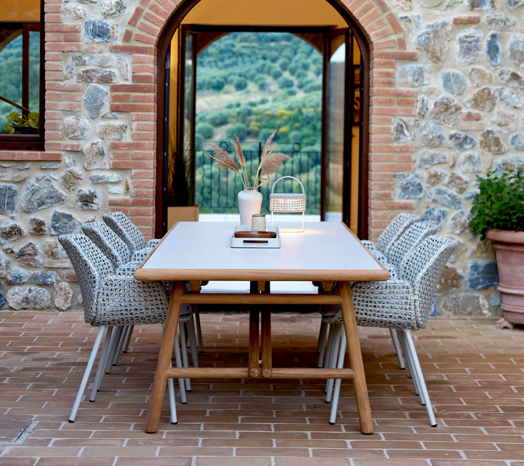 Una gran mesa de comedor rectangular con sillas tejidas, con un centro de bandeja de club y una lámpara de ilusión, contra un fondo de piedra.
