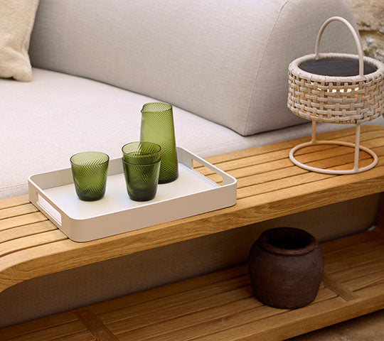 Una mesa de café de madera con una bandeja con cristalería verde, una cesta decorativa y una maceta de cerámica oscura.