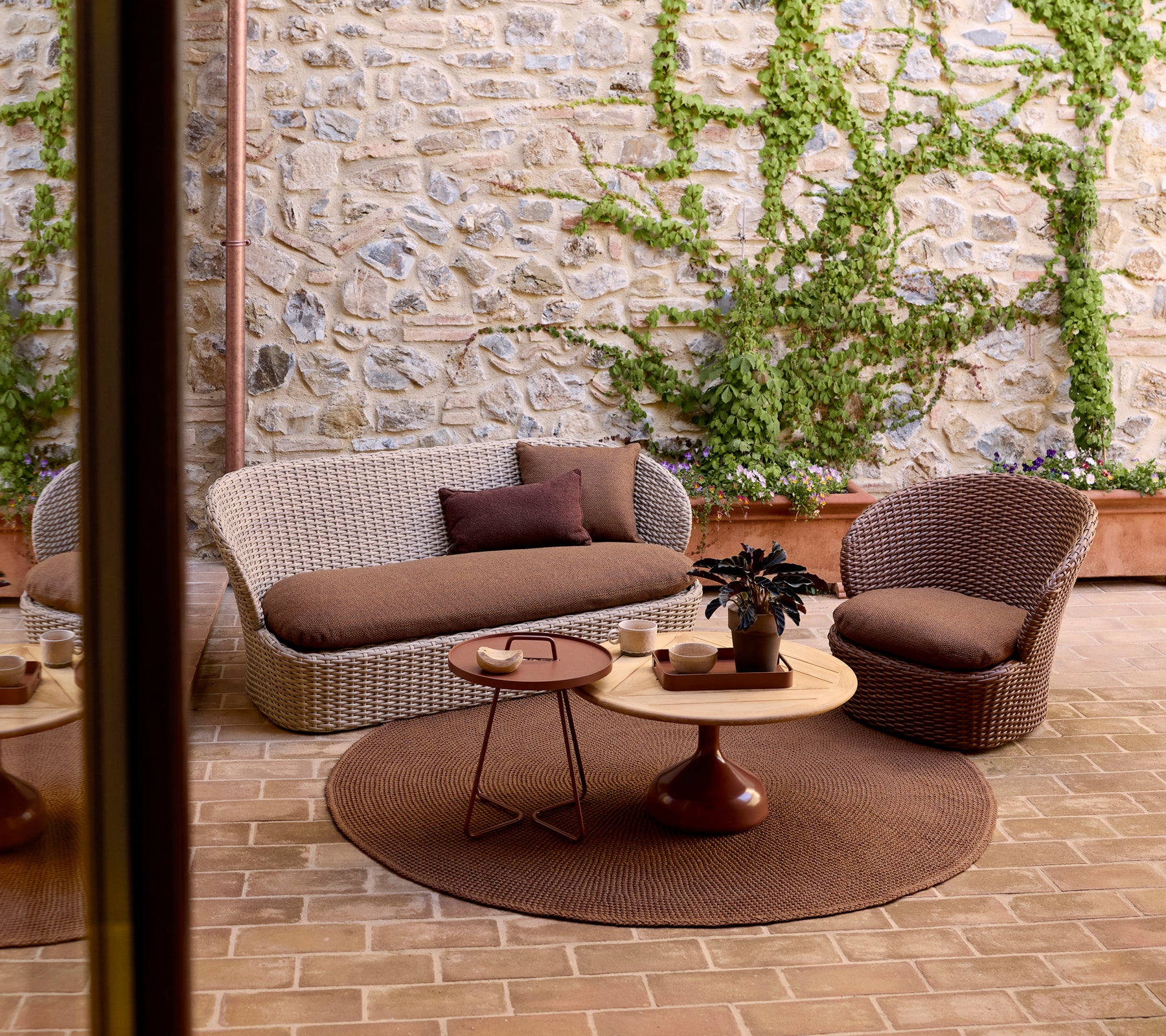 Área de lounge al aire libre acogedora con un sofá tejido, una silla, una mesa lateral redonda y vegetación contra una pared de piedra.