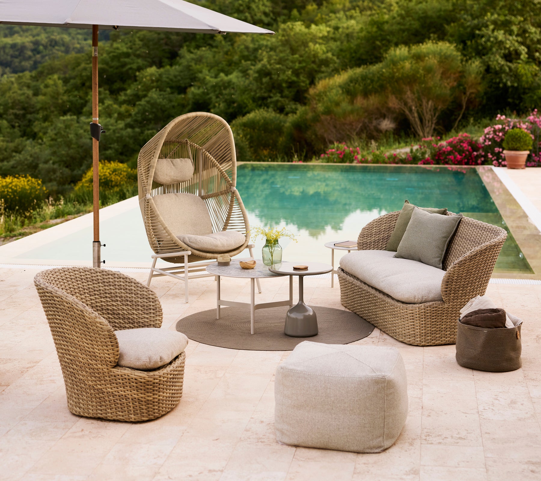 Configuración de salón costero con una silla Hive, dos sillas curvadas, un pouf Divine y una mesa pequeña junto a una piscina rodeada de vegetación.