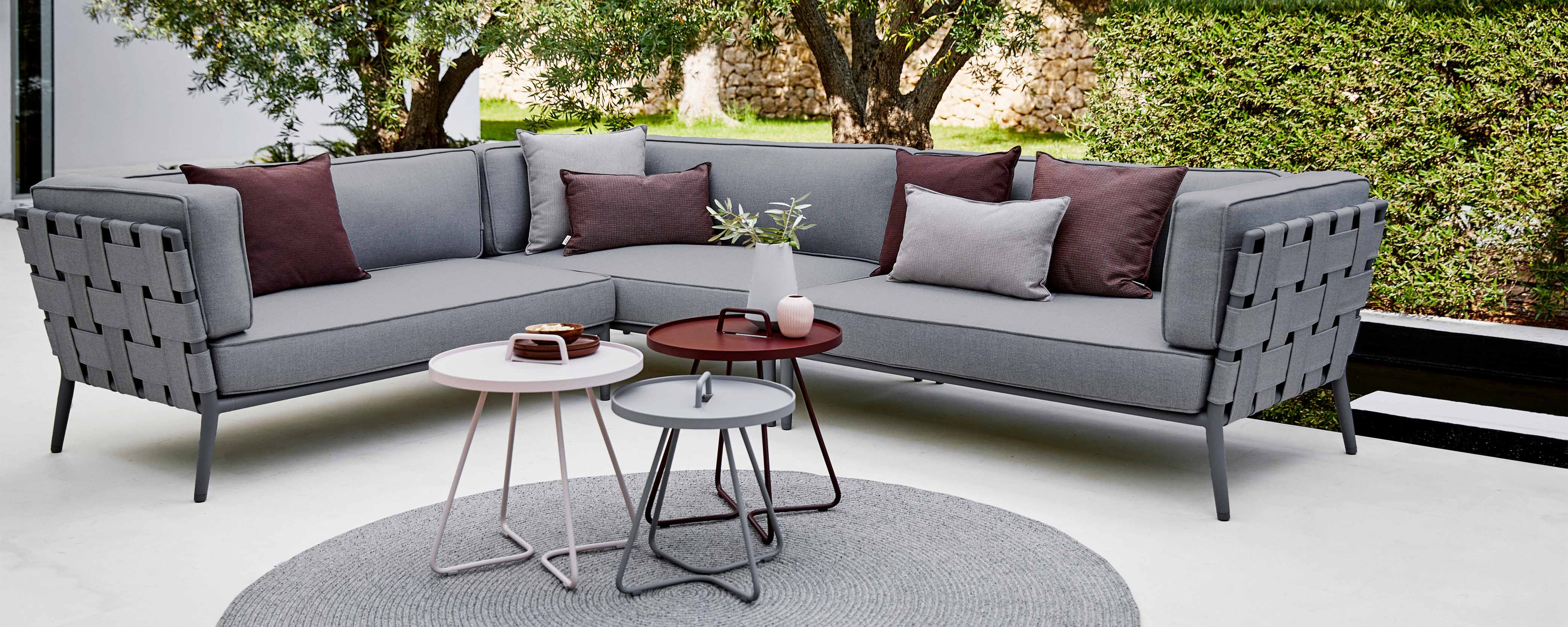 Sofá seccional cónico moderno en tela gris con cojines decorativos, acompañado de mesas auxiliares elegantes y una alfombra redonda.