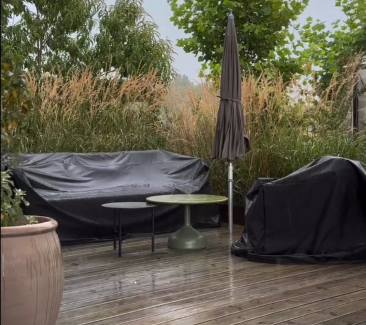 Espacio exterior con sofá negro, mesa verde y silla negra rodeada de vegetación.
