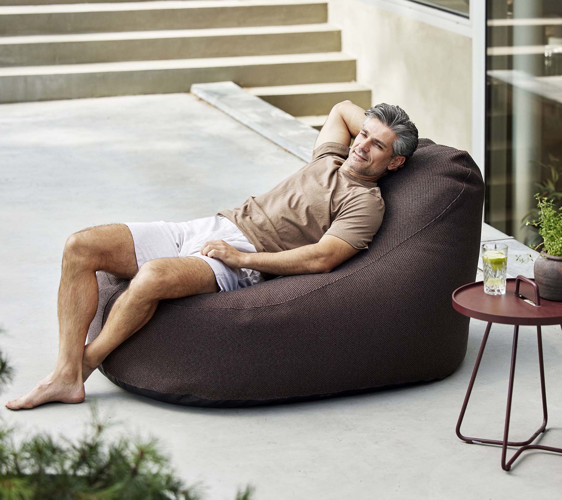 Un hombre se relaja en una cómoda silla de frijol, disfrutando de una bebida al lado de una mesa pequeña, rodeado de vegetación.