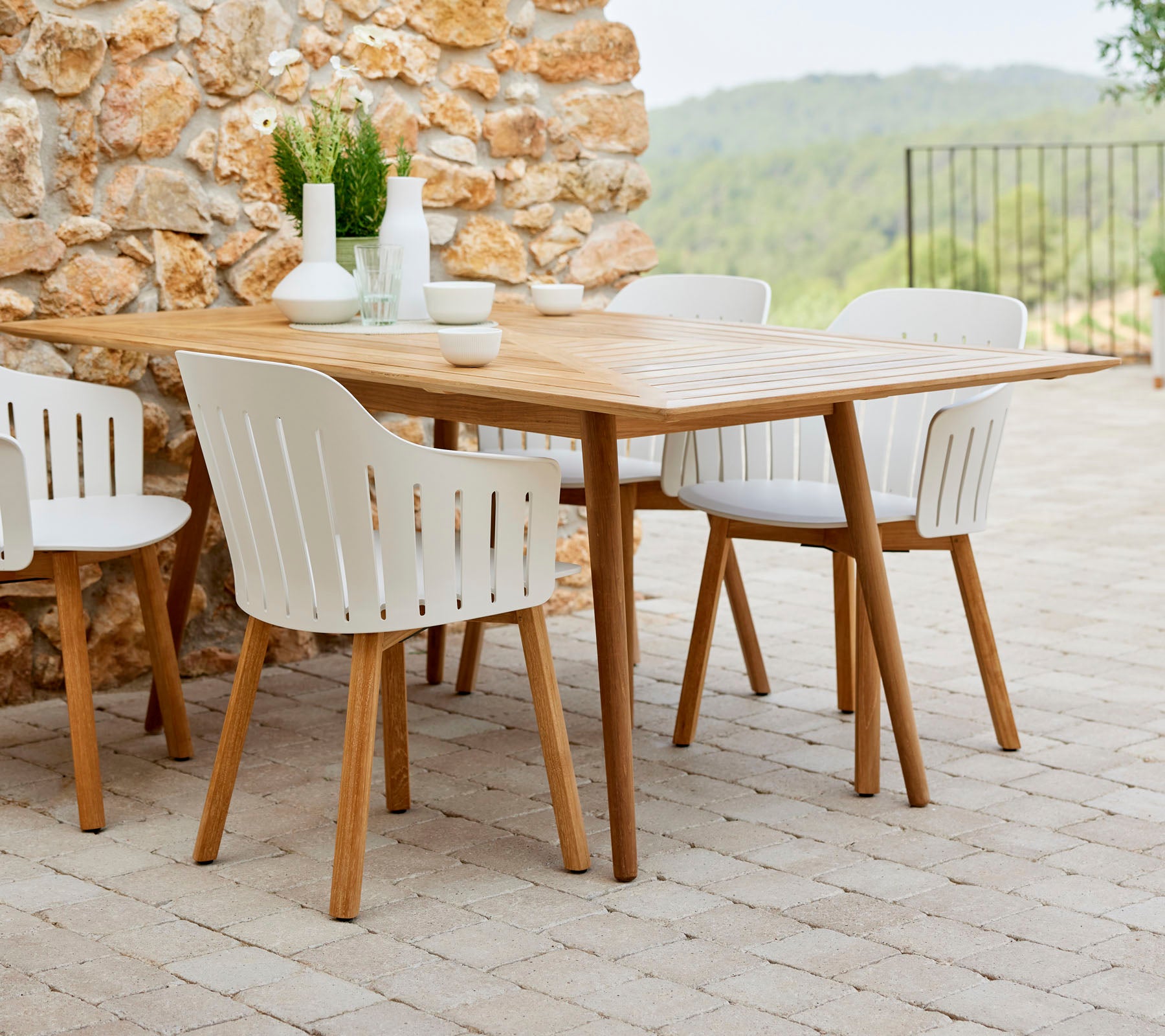 Una mesa de comedor de madera rodeada de sillas blancas, complementada con plantas decorativas y una vajilla simple, contra un fondo de piedra natural.