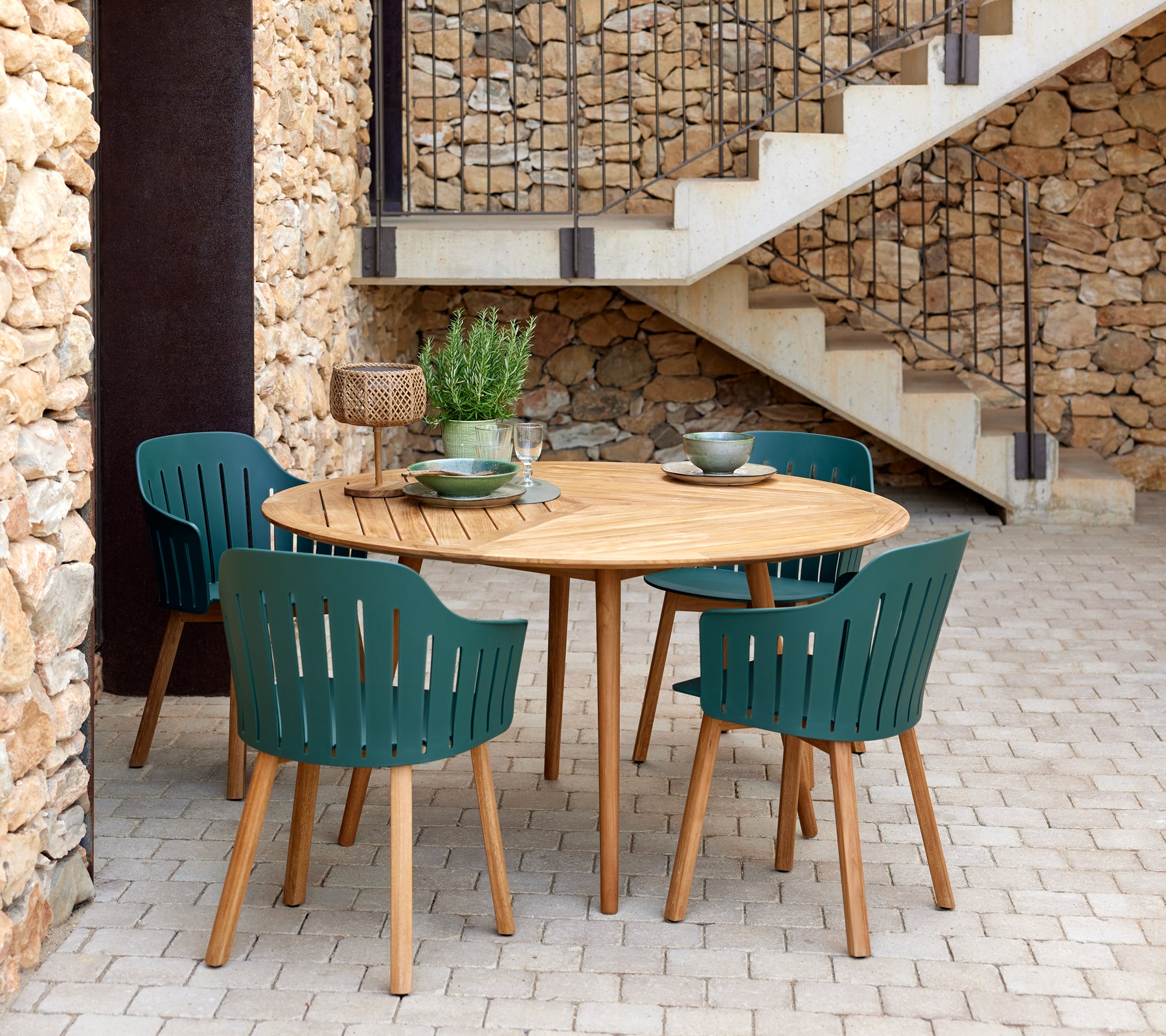 Una mesa redonda de madera rodeada de cuatro sillas verdes de listones, complementada con cuencos decorativos y una planta pequeña.
