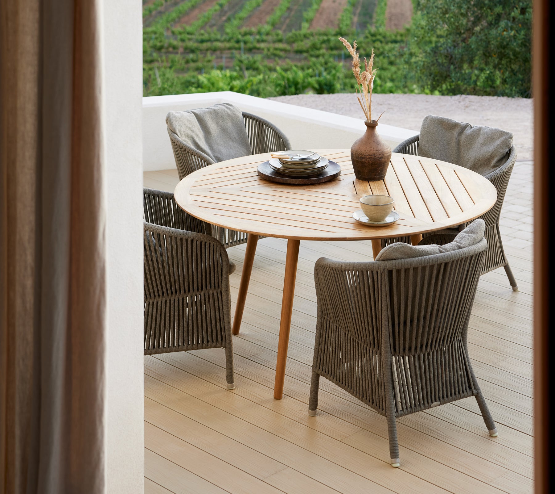 Una mesa redonda de madera rodeada de cuatro sillas tejidas, con un jarrón y un plato decorativo, frente a un paisaje pintoresco.