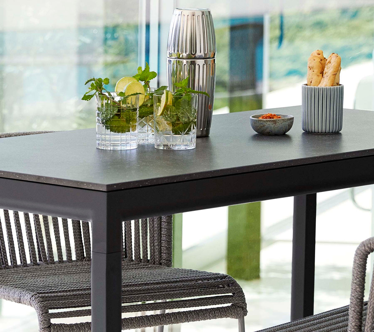 Una mesa moderna dispuesta con plantas en jarrones de vidrio, un recipiente decorativo y un plato de aperitivos, rodeada de sillas elegantes.