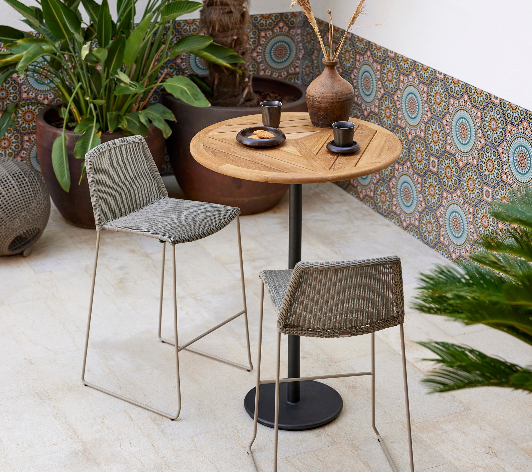 Un elegante juego de bistró con una mesa redonda de madera y dos taburetes trenzados, acompañado de plantas decorativas y vajilla moderna.