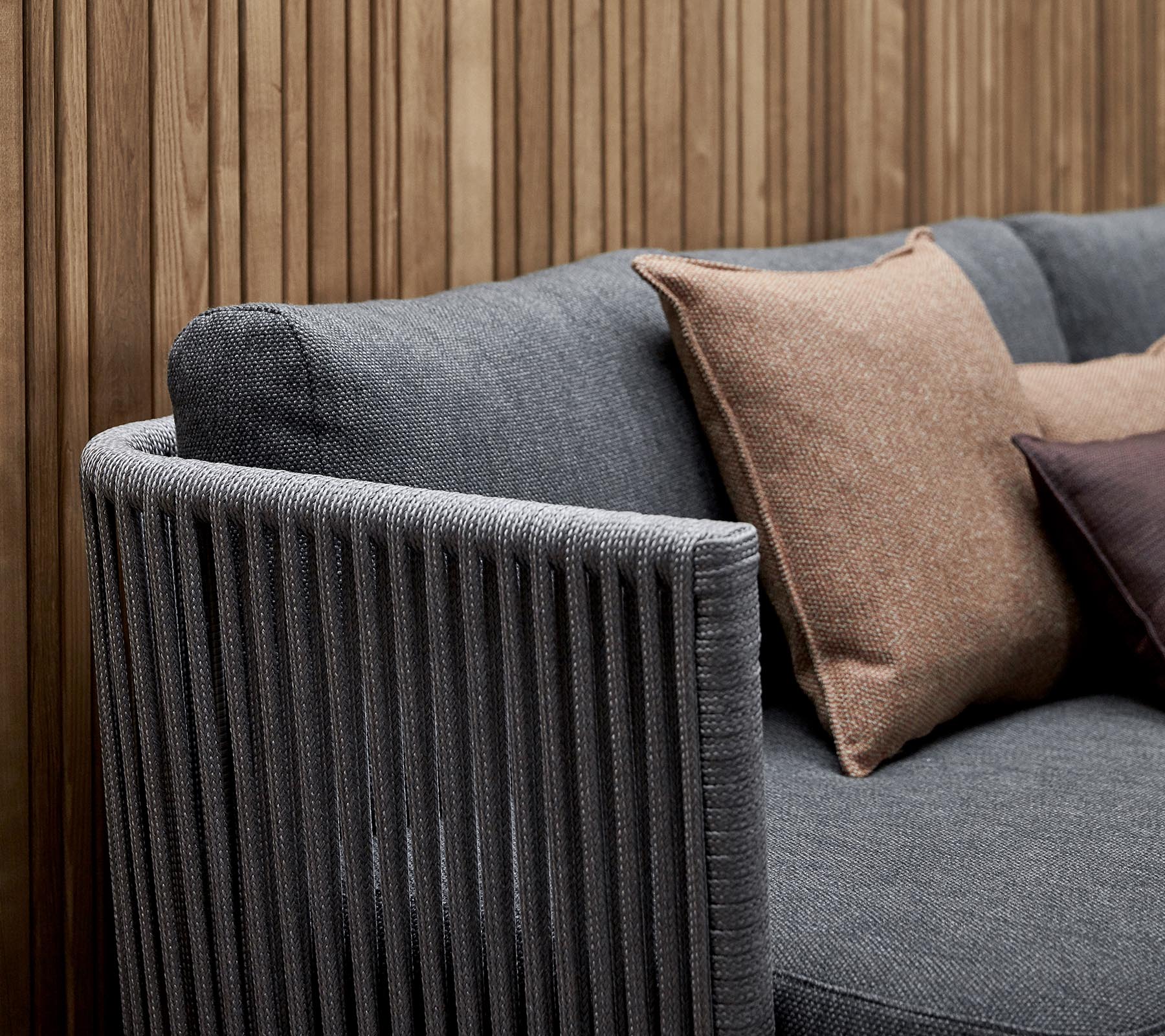 Sofá elegante con un tejido gris texturizado y cojines decorativos en tonos tierra, y un diseño de reposabrazos a rayas verticales.