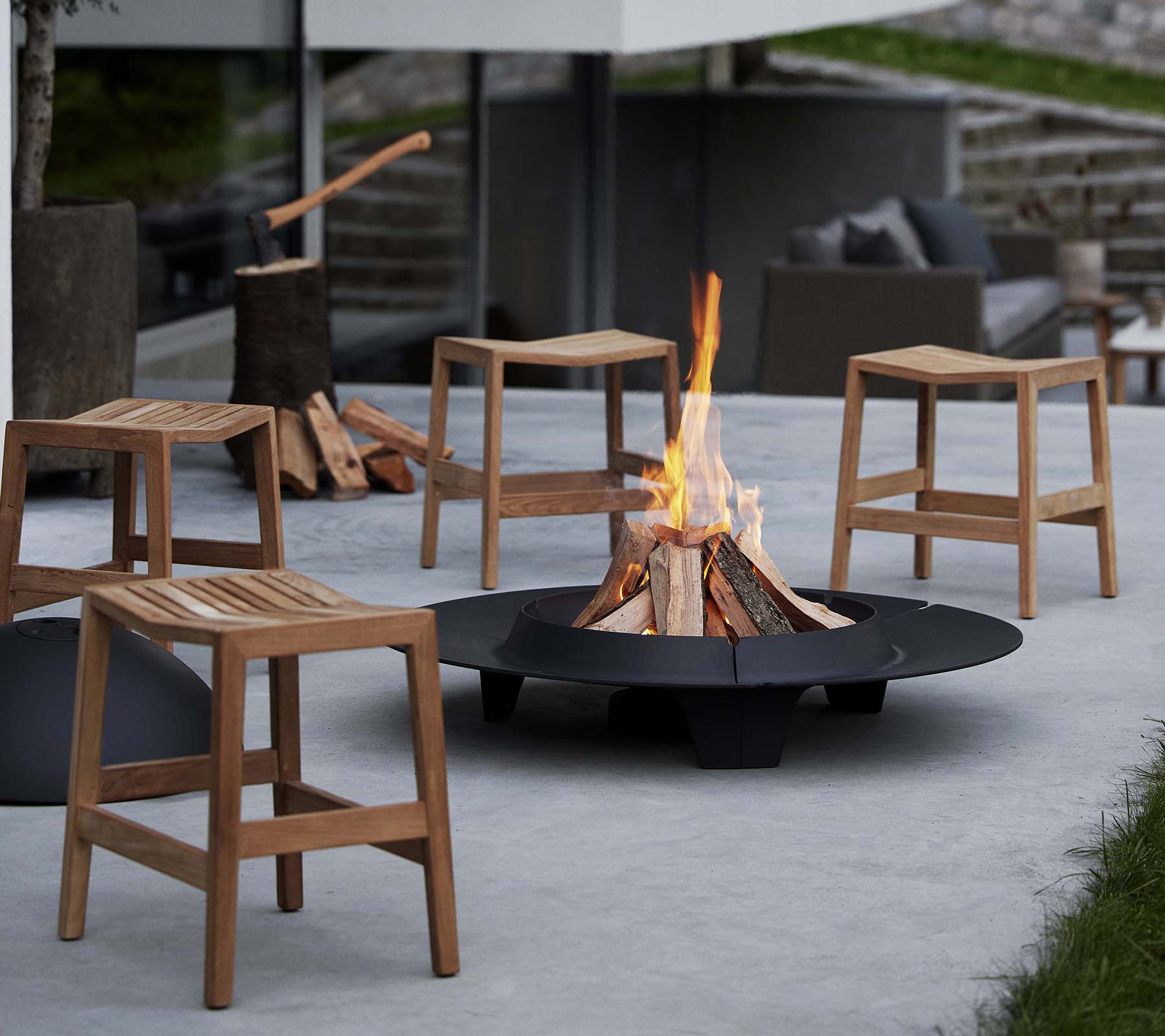 Taburetes de madera plegables rodean un fogón circular de brasas, con un fuego ardiente en el centro y troncos apilados cerca.