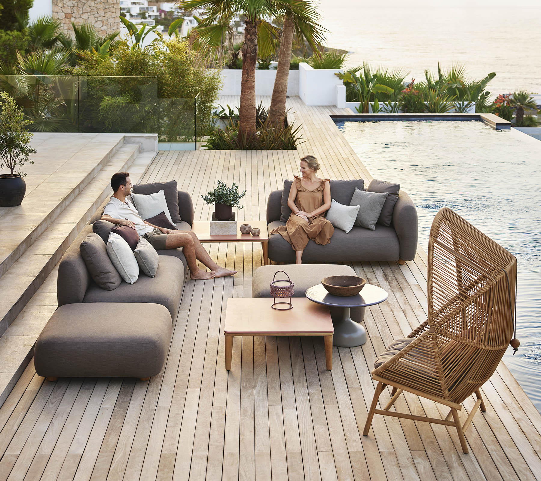 Dos personas se relajan en asientos modernos junto a una piscina, rodeados de vegetación, con una mesa de madera y plantas decorativas cerca.