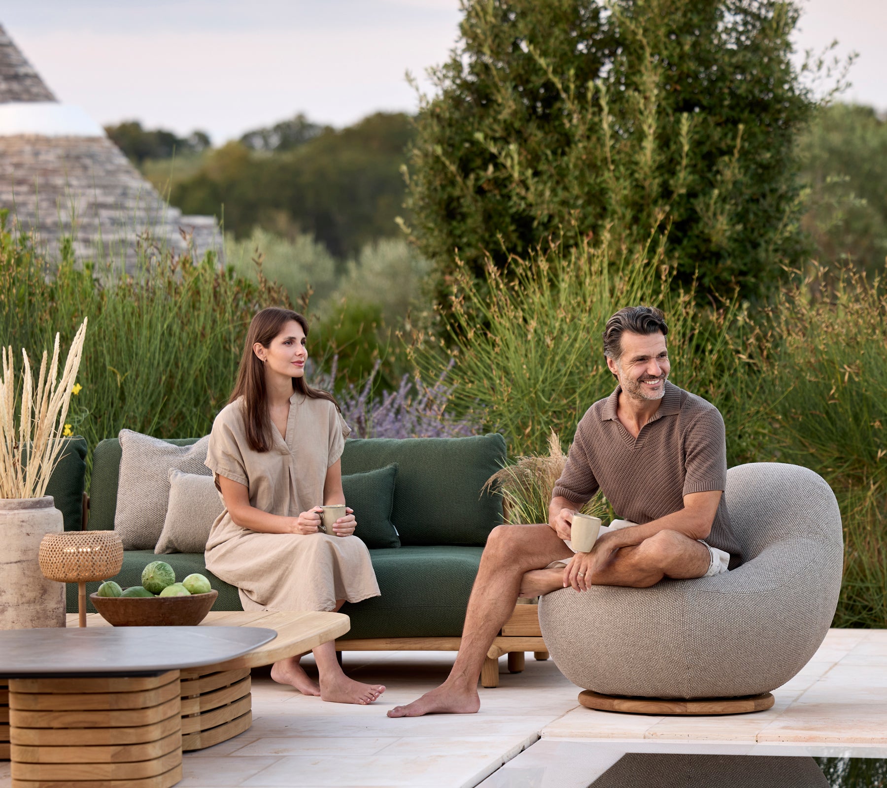 Sala moderna con sofá verde, silla redonda gris y mesa marrón rodeada de vegetación.
