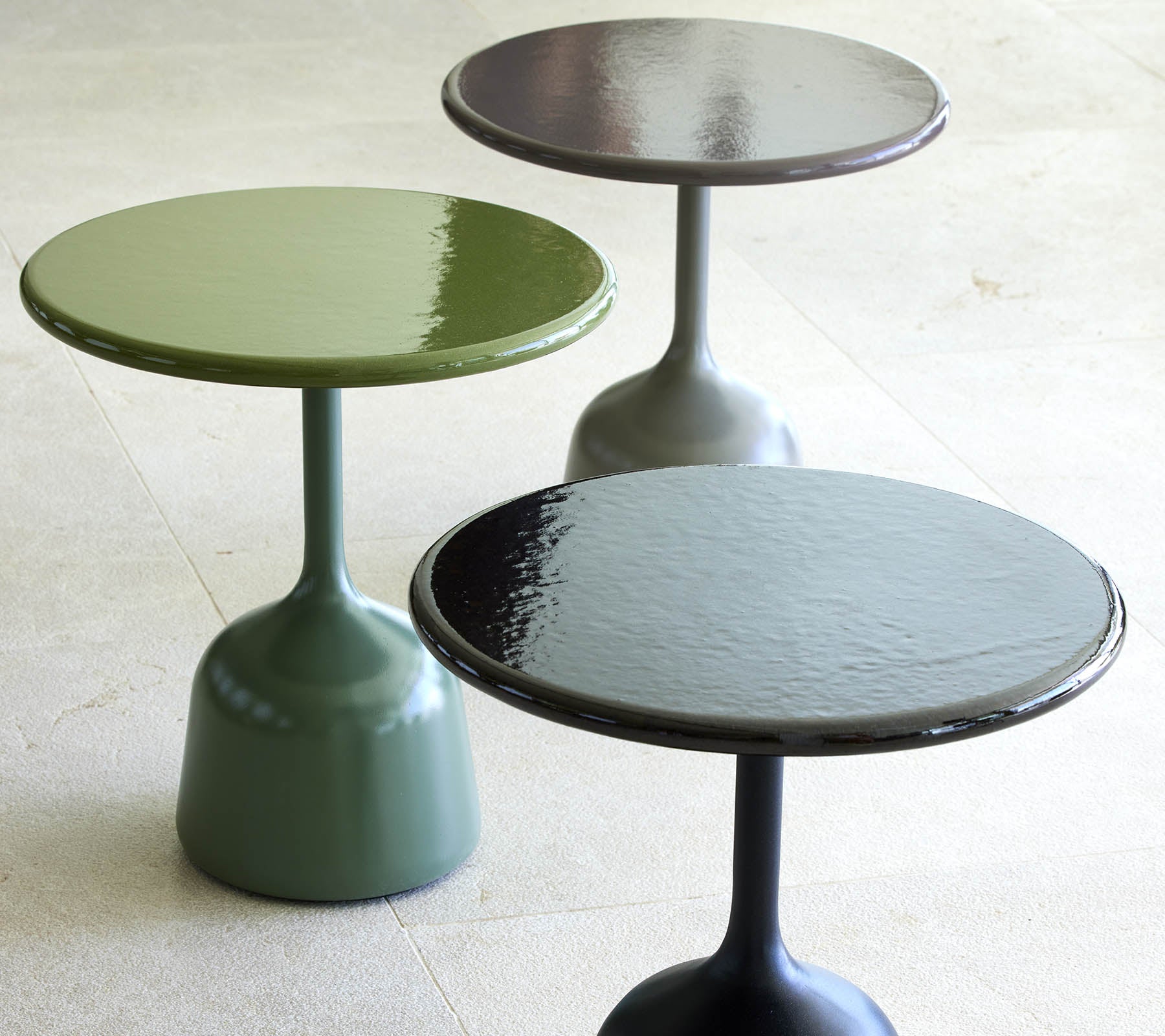Tres mesas de café lacadas en verde, negro y gris oscuro, cada una con una parte superior redonda y una base esbelta y curvada.