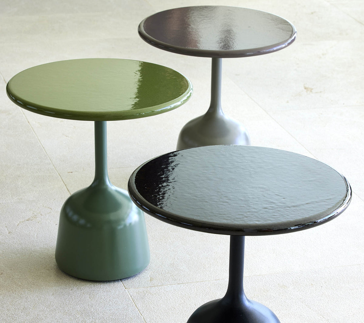Tres mesas de café lacadas en verde, negro y gris oscuro, cada una con una parte superior redonda y una base esbelta y curvada.