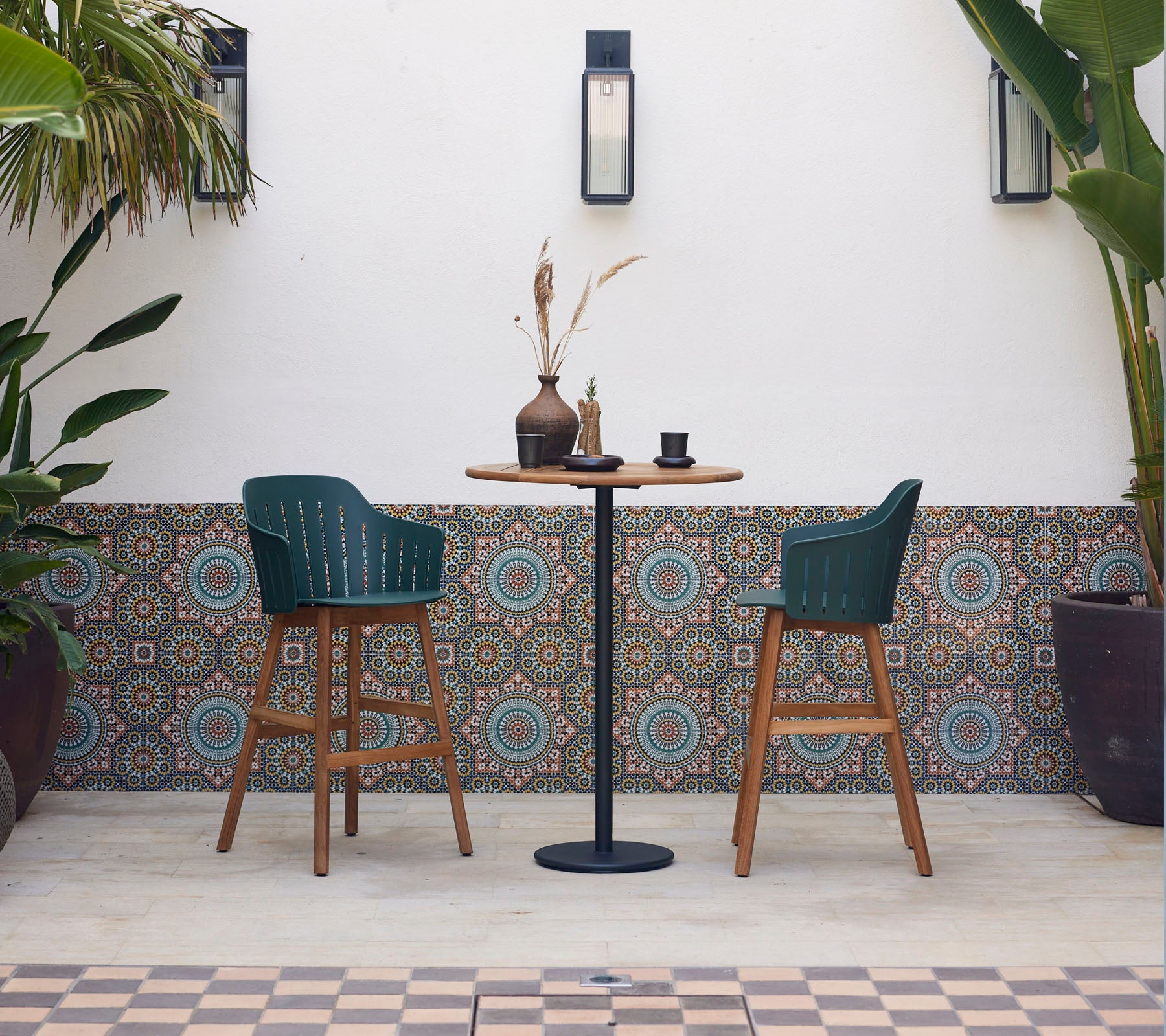 Una mesa de bar elegante con dos sillas verdes, decorada con un jarrón y una taza, rodeada de azulejos decorados y plantas exuberantes.