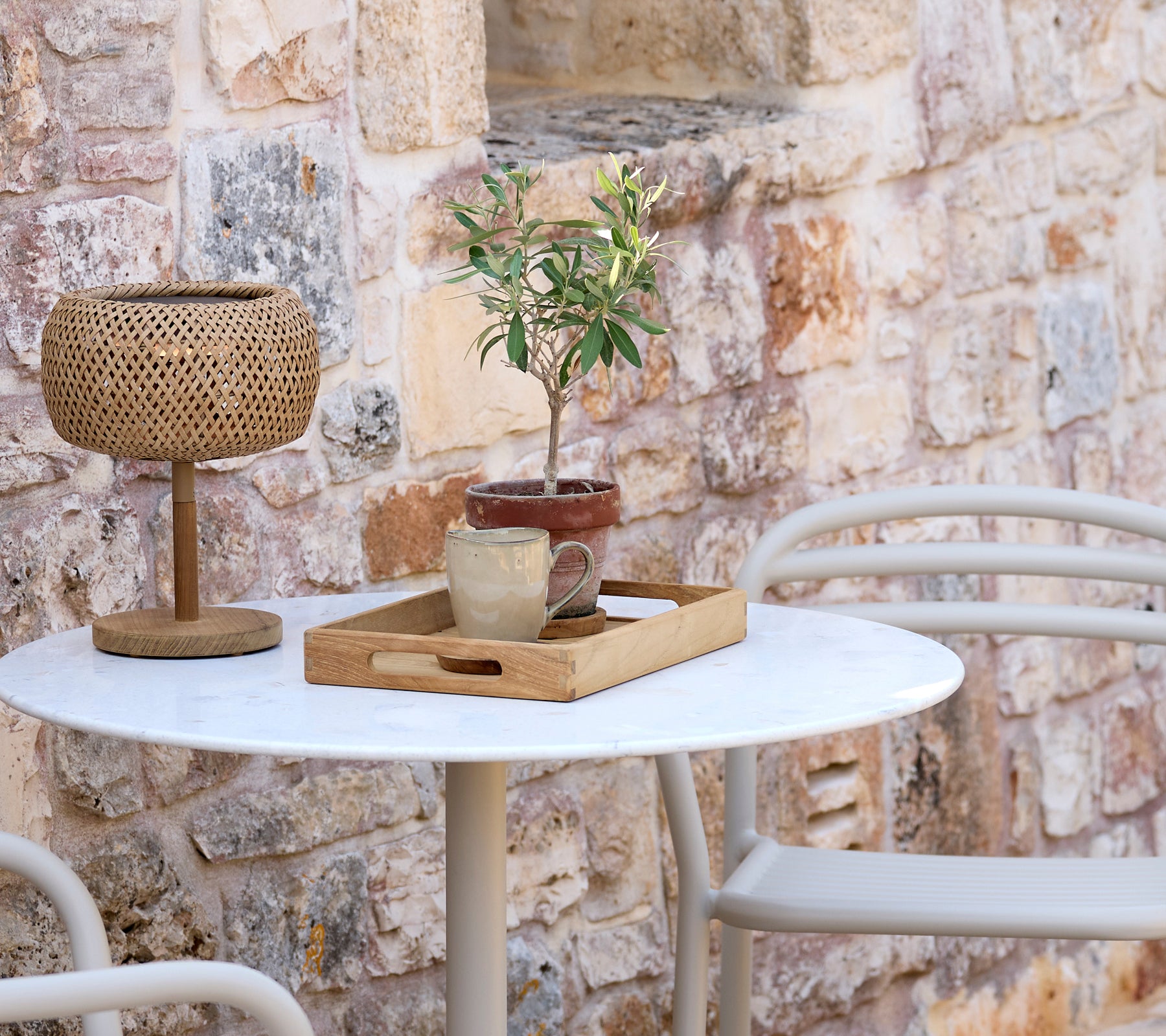 Montaje moderno al aire libre con una mesa blanca, una lámpara tejida, una bandeja de madera y una planta en maceta.