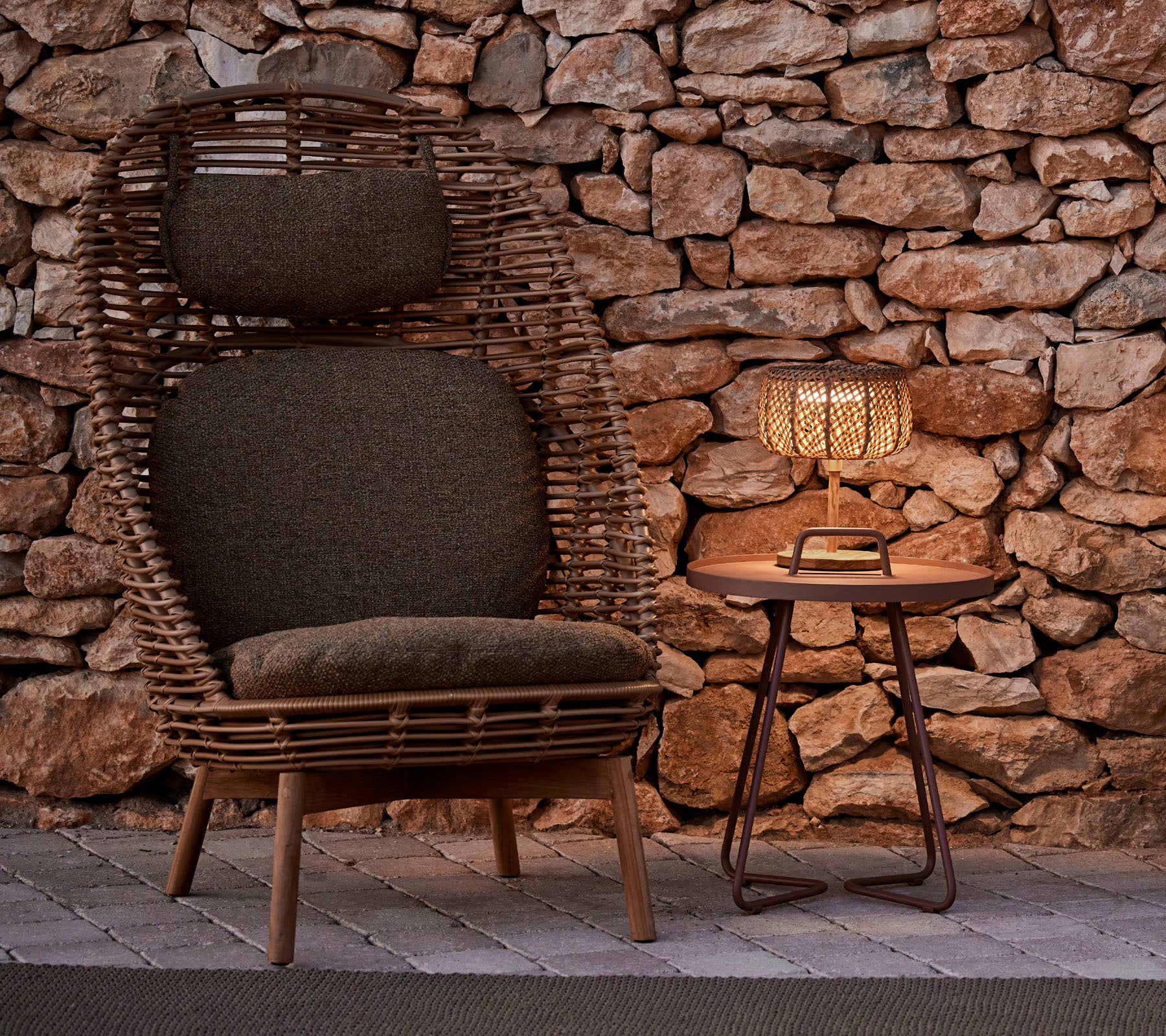 Una silla tejida elegante con cojines al lado de una mesa de madera y una lámpara brillante, contra un fondo de piedra rústica.