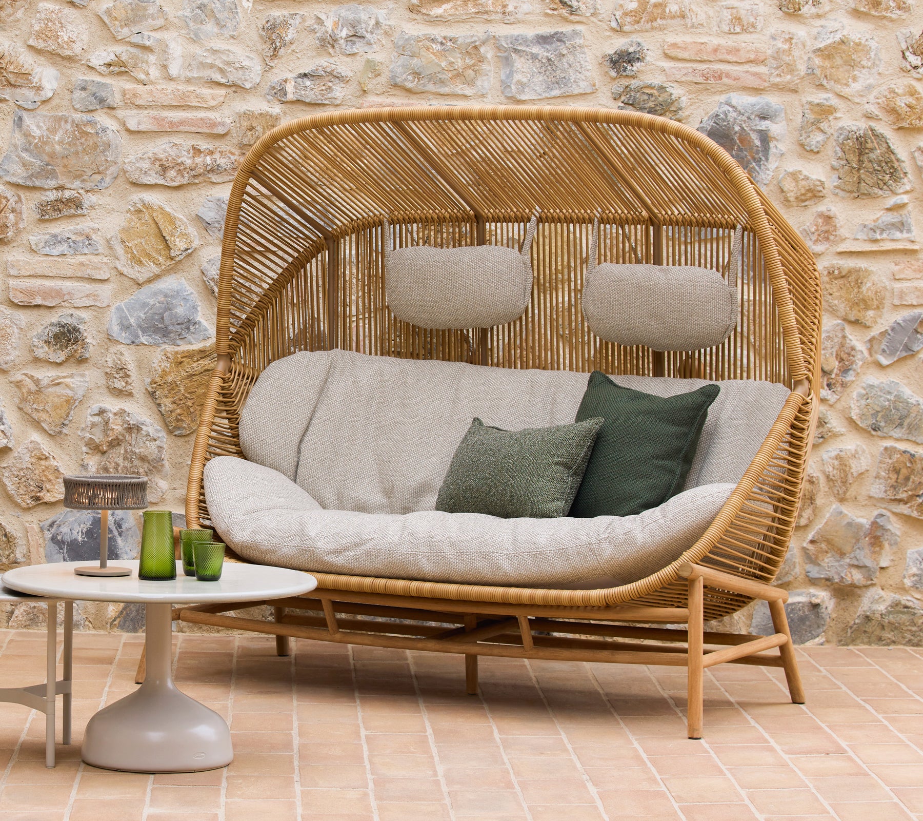 Sofá Hive con cojines mullidos y acentos verdes junto a mesas de café Glaze, con un fondo de piedra natural.