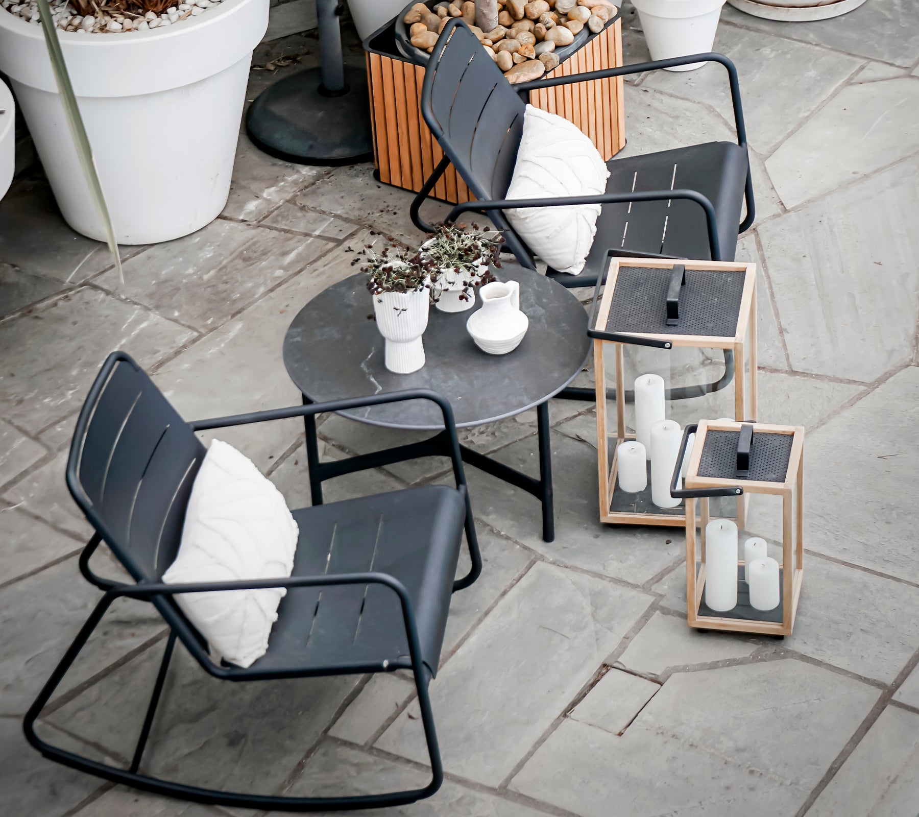 Dos sillas mecedoras modernas y una mesa redonda con plantas decorativas y faroles, dispuestas sobre una superficie de piedra texturizada.