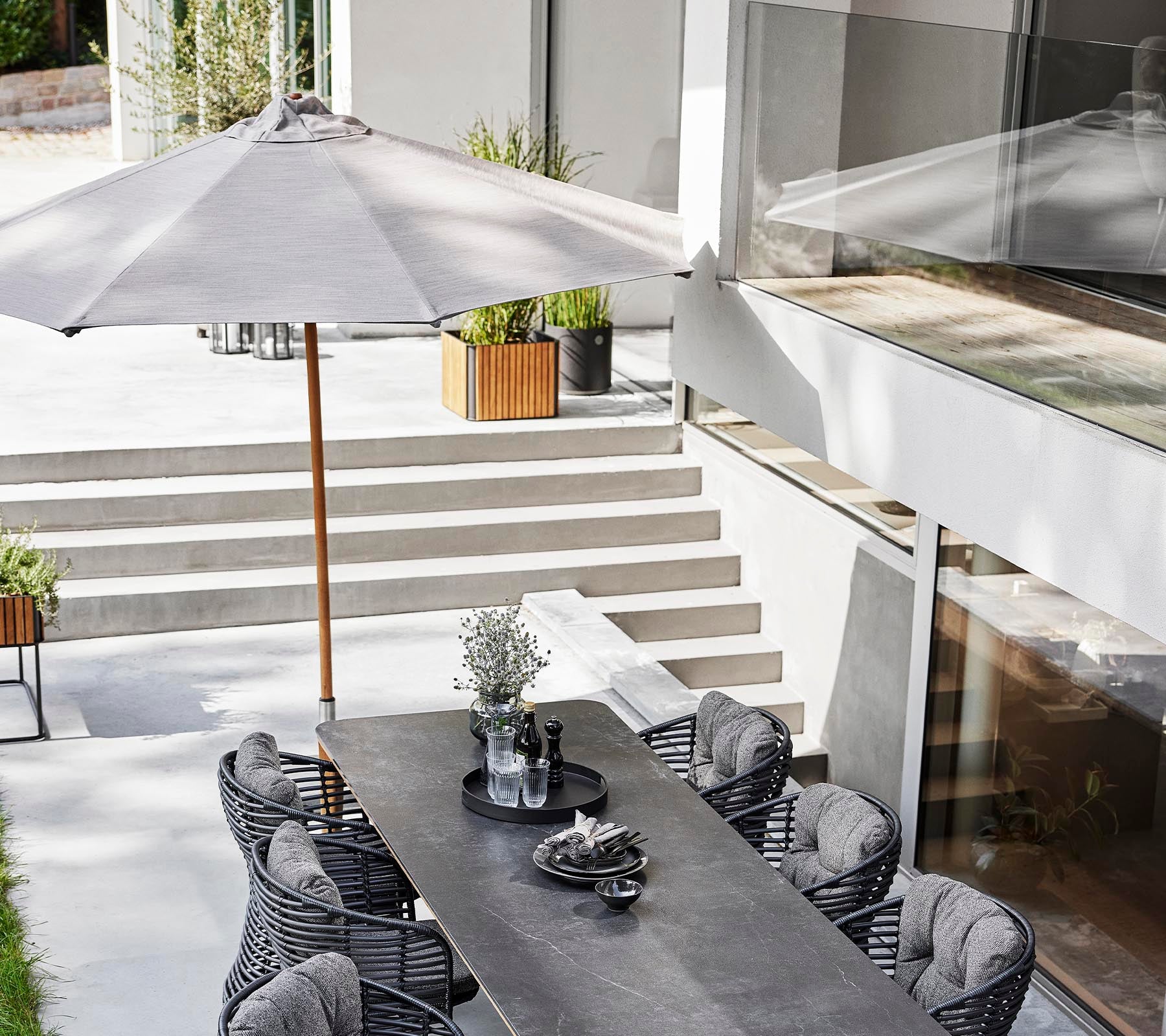 Una elegante configuración de comedor al aire libre con una mesa larga, sillas de mimbre y un parasol clásico para sombra.