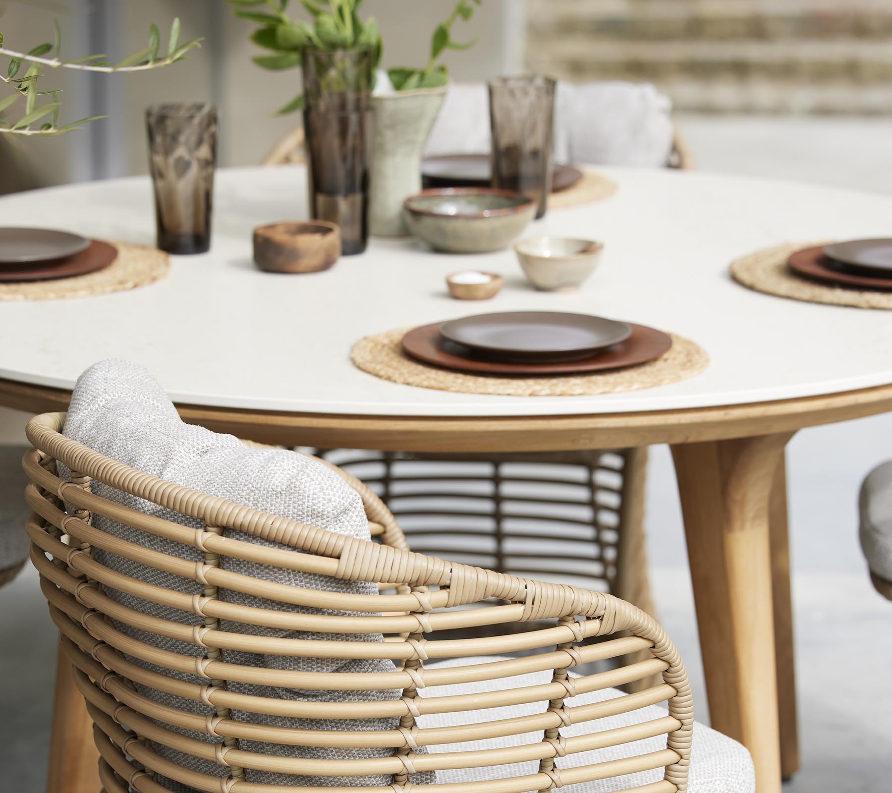 Un elegante juego de mesa de comedor con platos, cuencos y copas decorativas, rodeado de sillas trenzadas y elementos naturales.