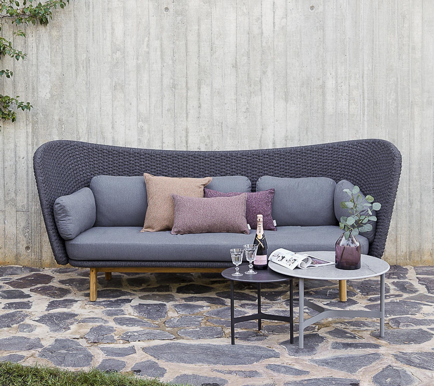 Un sofá moderno en gris con cojines decorativos, acompañado de una mesa pequeña con bebidas y un jarrón, situado contra una pared texturizada.